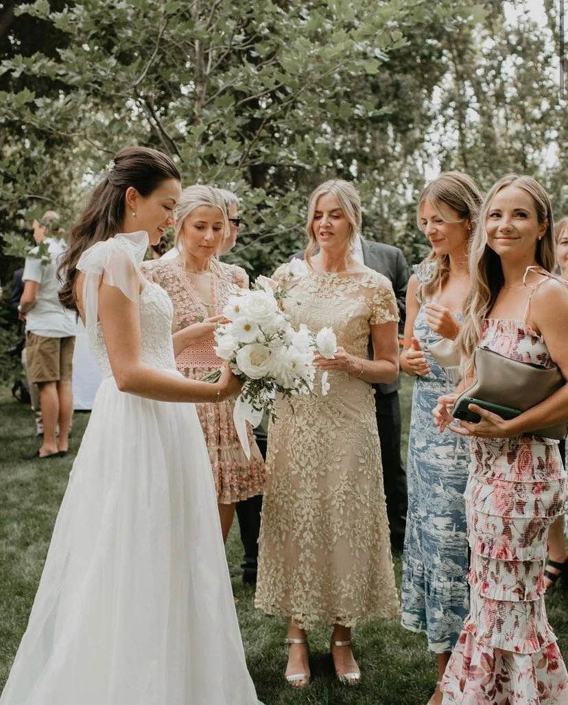 A bride holding a bouquet of white flowers surrounded by women at an outdoor wedding reception.