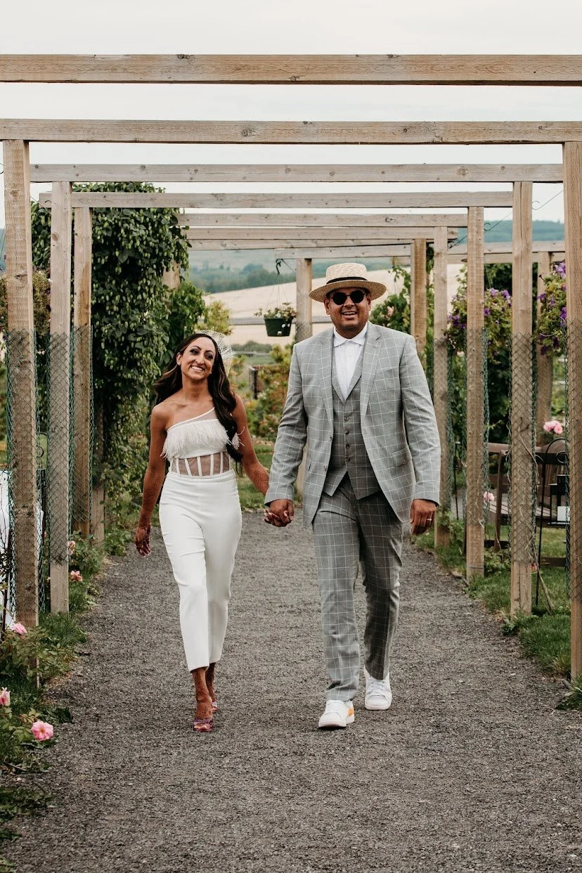 Couple walking hand in hand through a garden aisle, dressed in wedding attire, smiling, with greenery and flowers on either side.