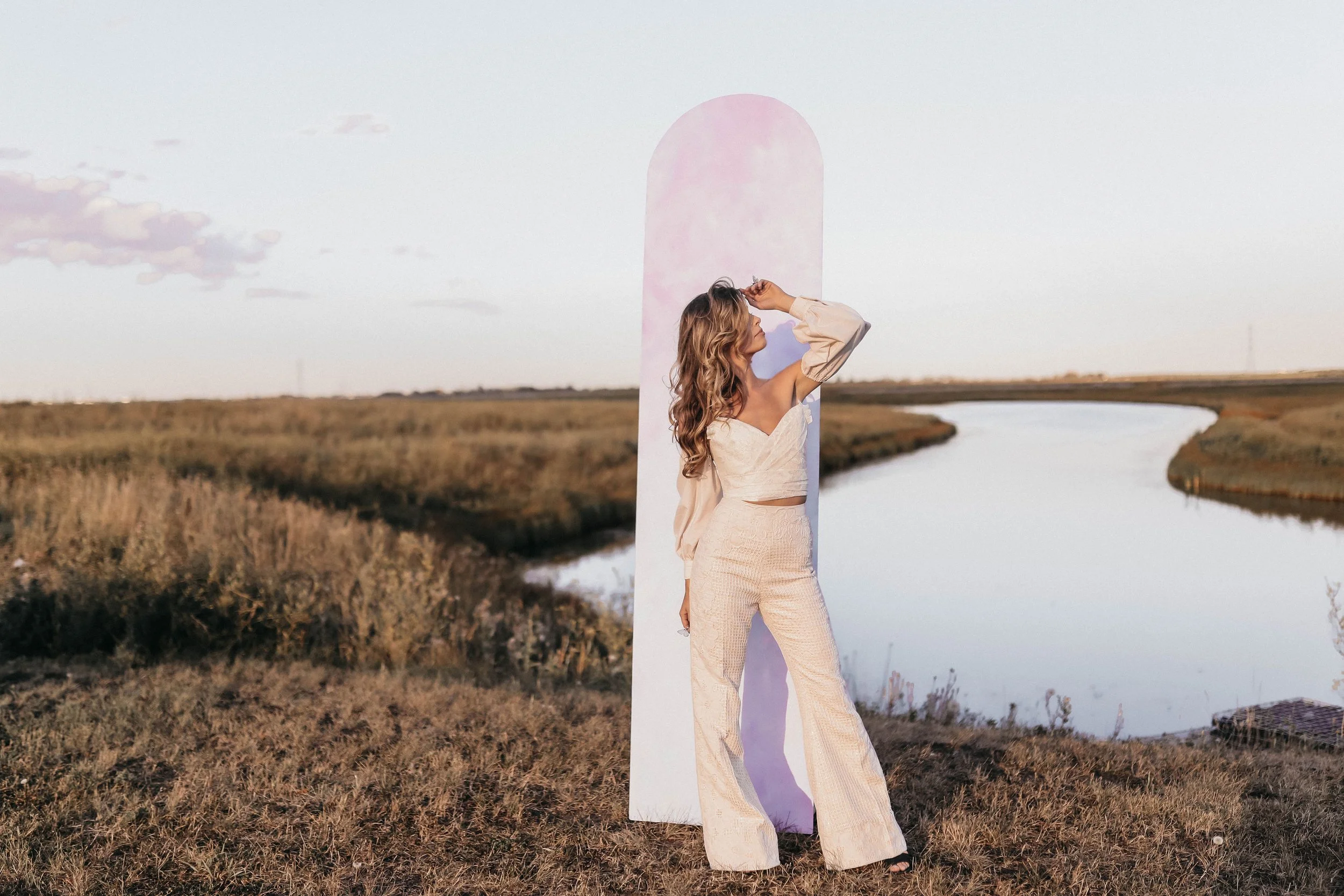 A woman with long wavy blonde hair stands in an open field near a river, posing with one hand on her forehead. She wears a beige off-the-shoulder top and matching wide-leg pants. A tall, pinkish-white rectangular structure is behind her. The sky is c