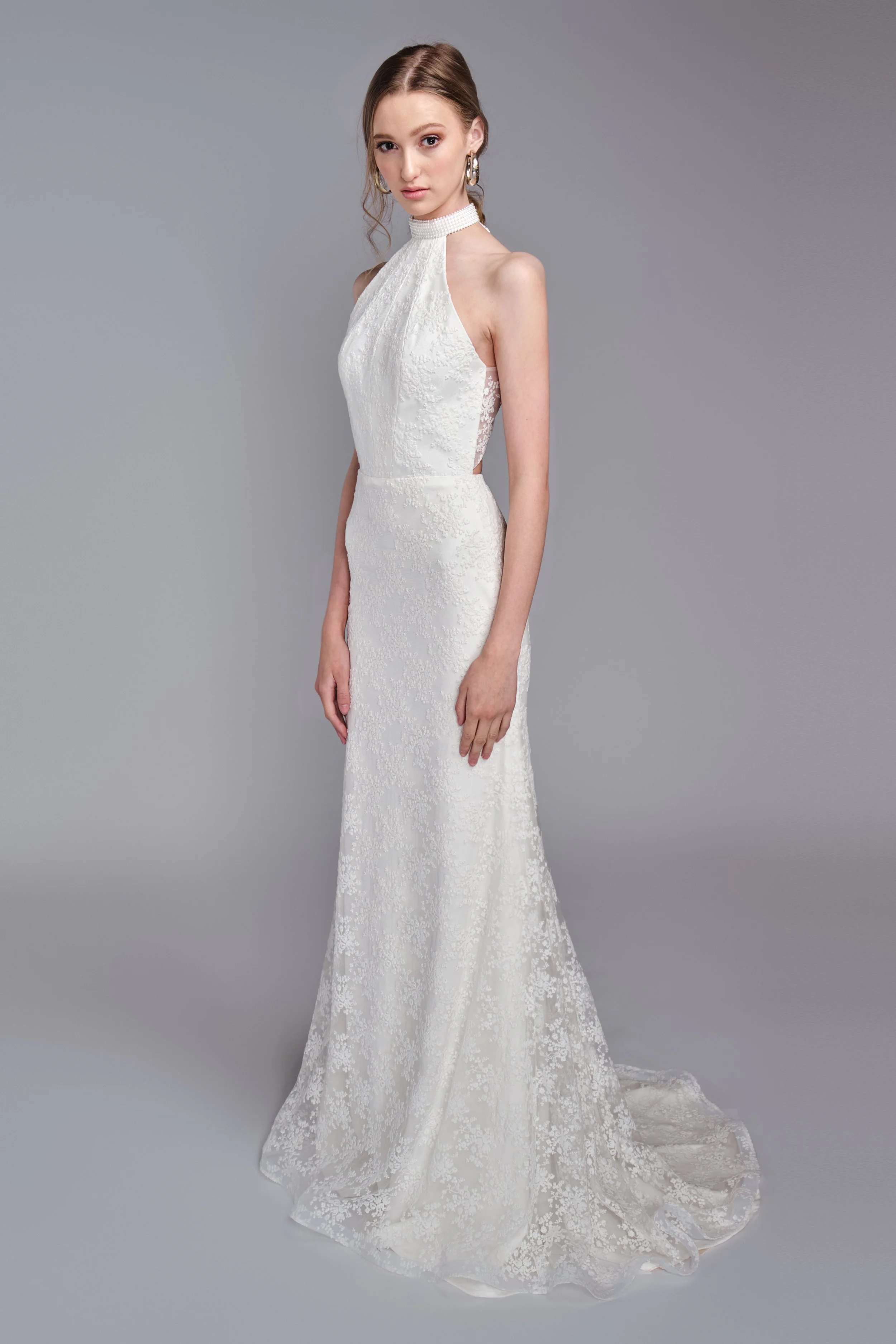 A woman in a white lace wedding dress with a high neck, side cutouts, and a train, standing against a plain gray background.