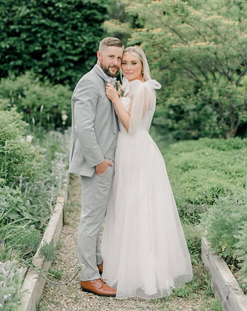 A couple dressed in wedding attire standing outdoors on a garden path, with lush greenery in the background.