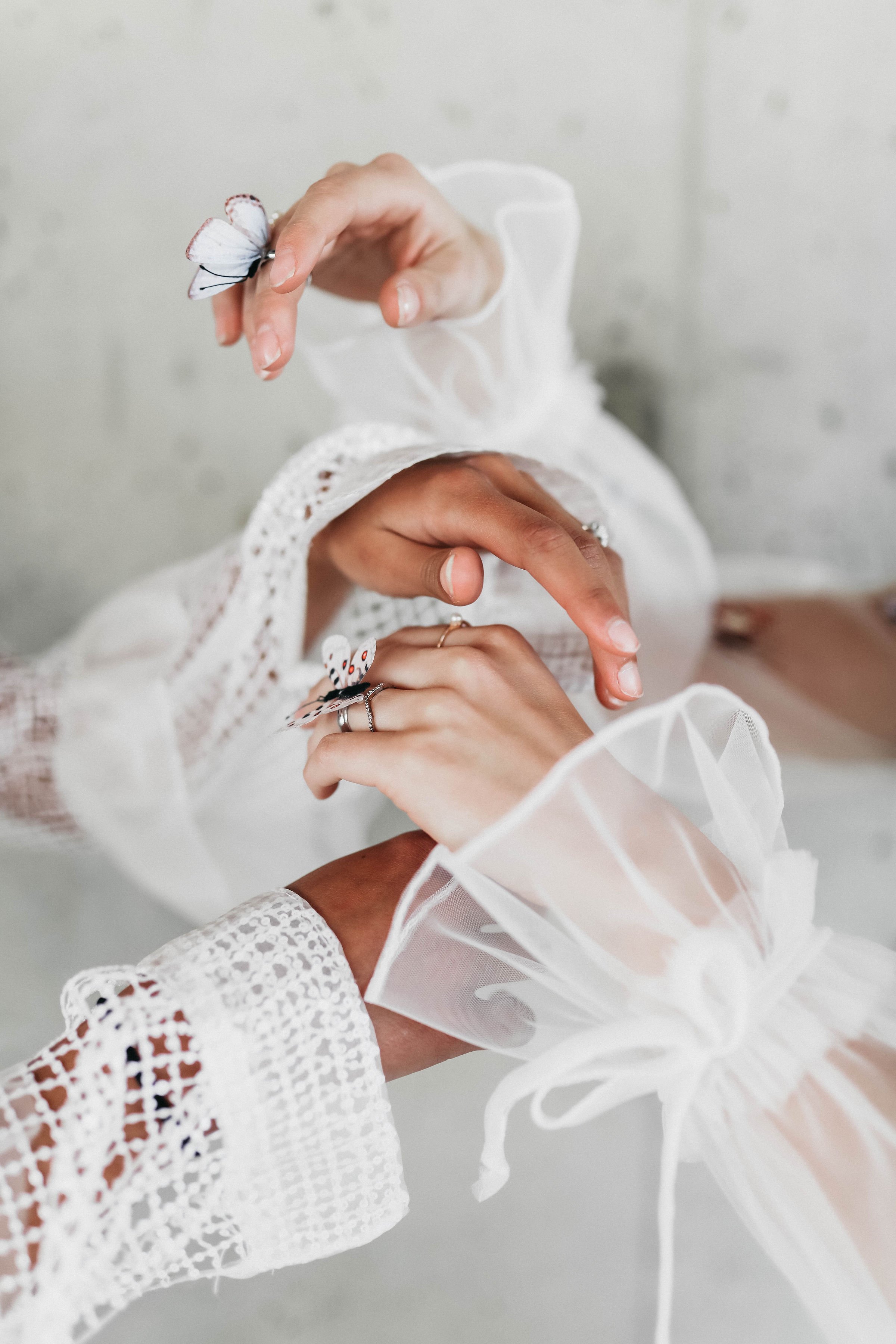 Close-up of hands wearing rings and lace cuffs, gently holding two white butterflies with black and red markings, against a soft, neutral background.