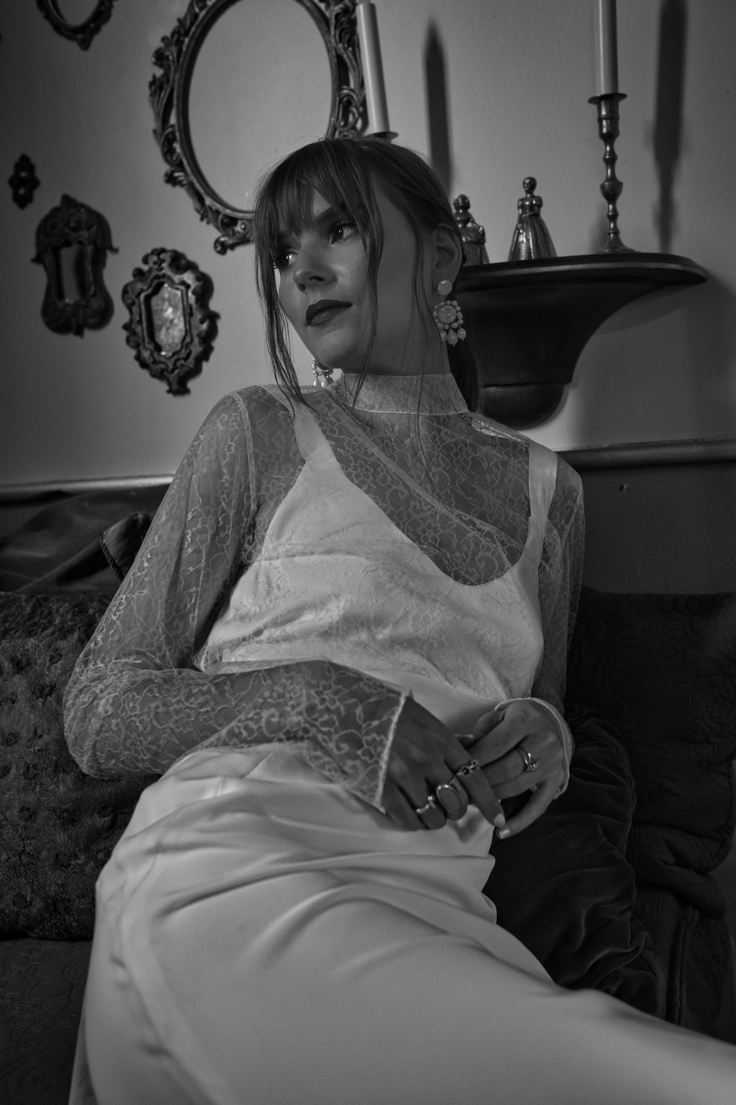 A woman with dark hair and earrings sitting on a couch in a room decorated with ornate mirrors and a shelf with candlesticks.