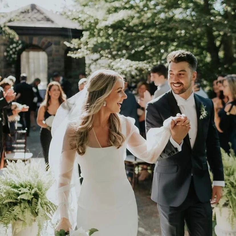 A bride and groom are dancing at their wedding reception outside, surrounded by guests and greenery.