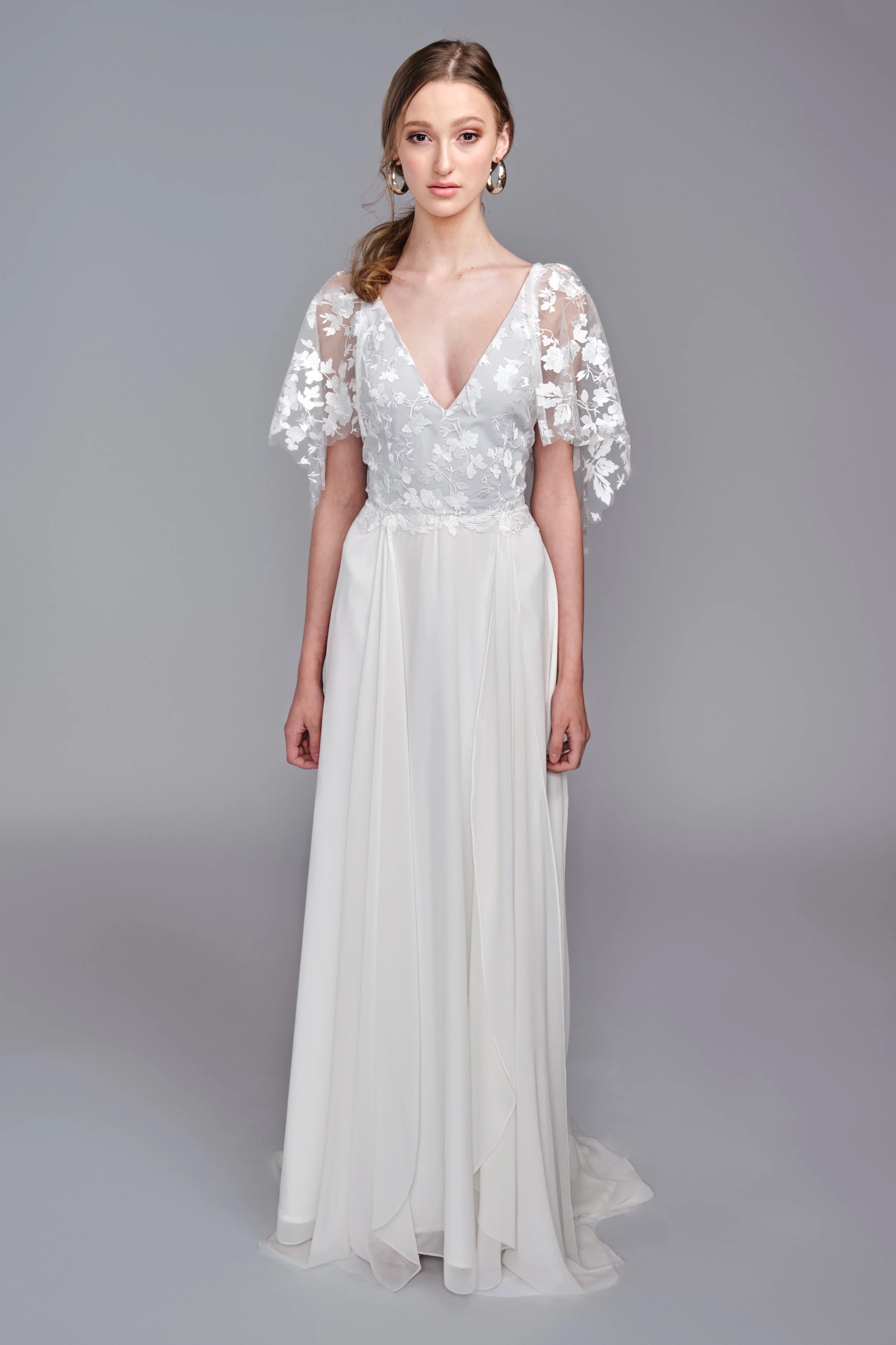 A young woman with light brown hair wearing a white dress with lace floral patterns on the bodice and sheer lace sleeves, standing against a plain gray background.