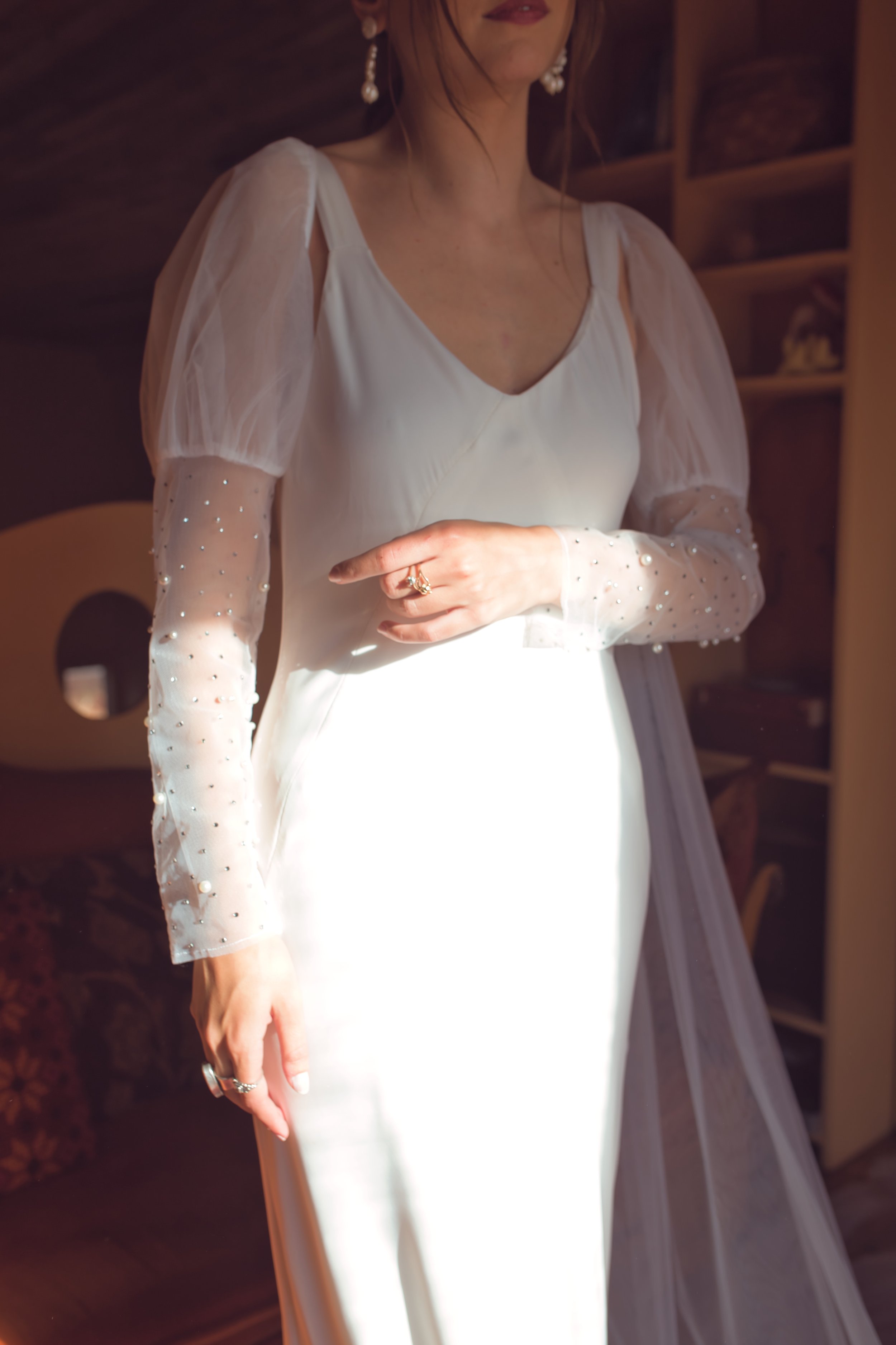 Close-up of a woman in a white dress with sheer, embellished sleeves, wearing pearl earrings and rings, standing indoors with wooden shelves in the background.