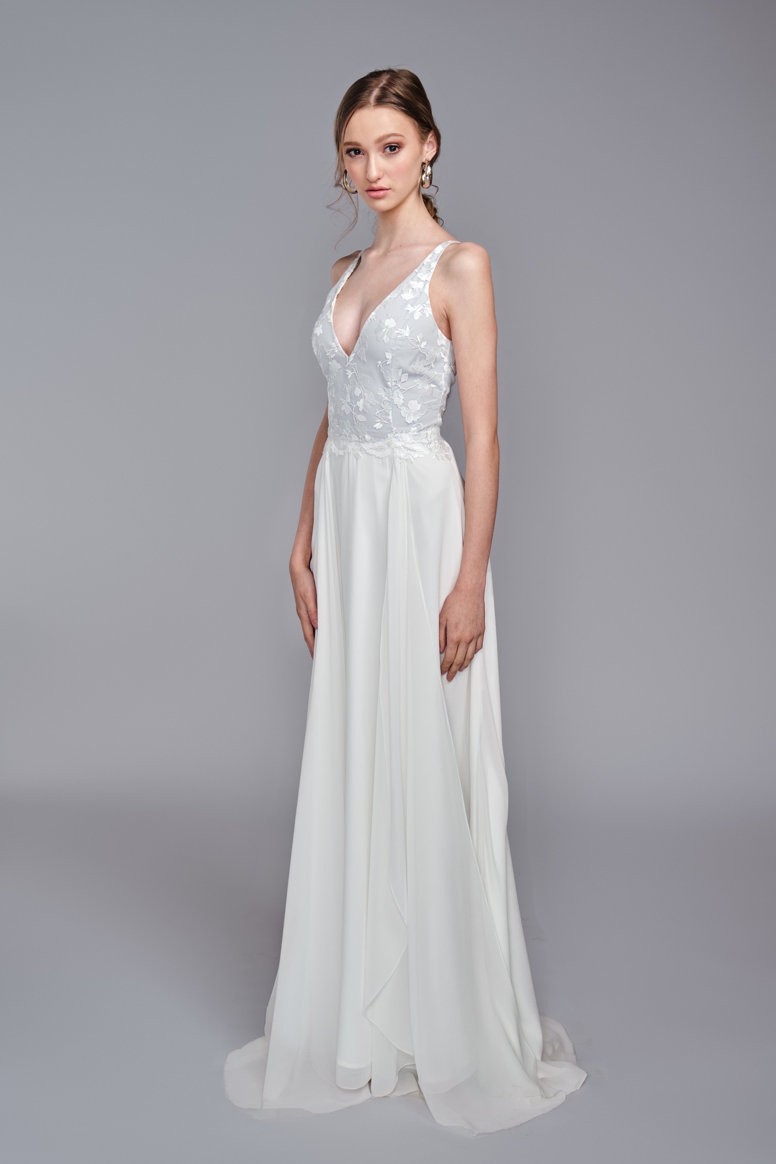 A young woman with light skin and brown hair in a low ponytail wearing a white wedding gown with a V-neck neckline, lace bodice with floral pattern, and flowing skirt, standing against a plain gray background.