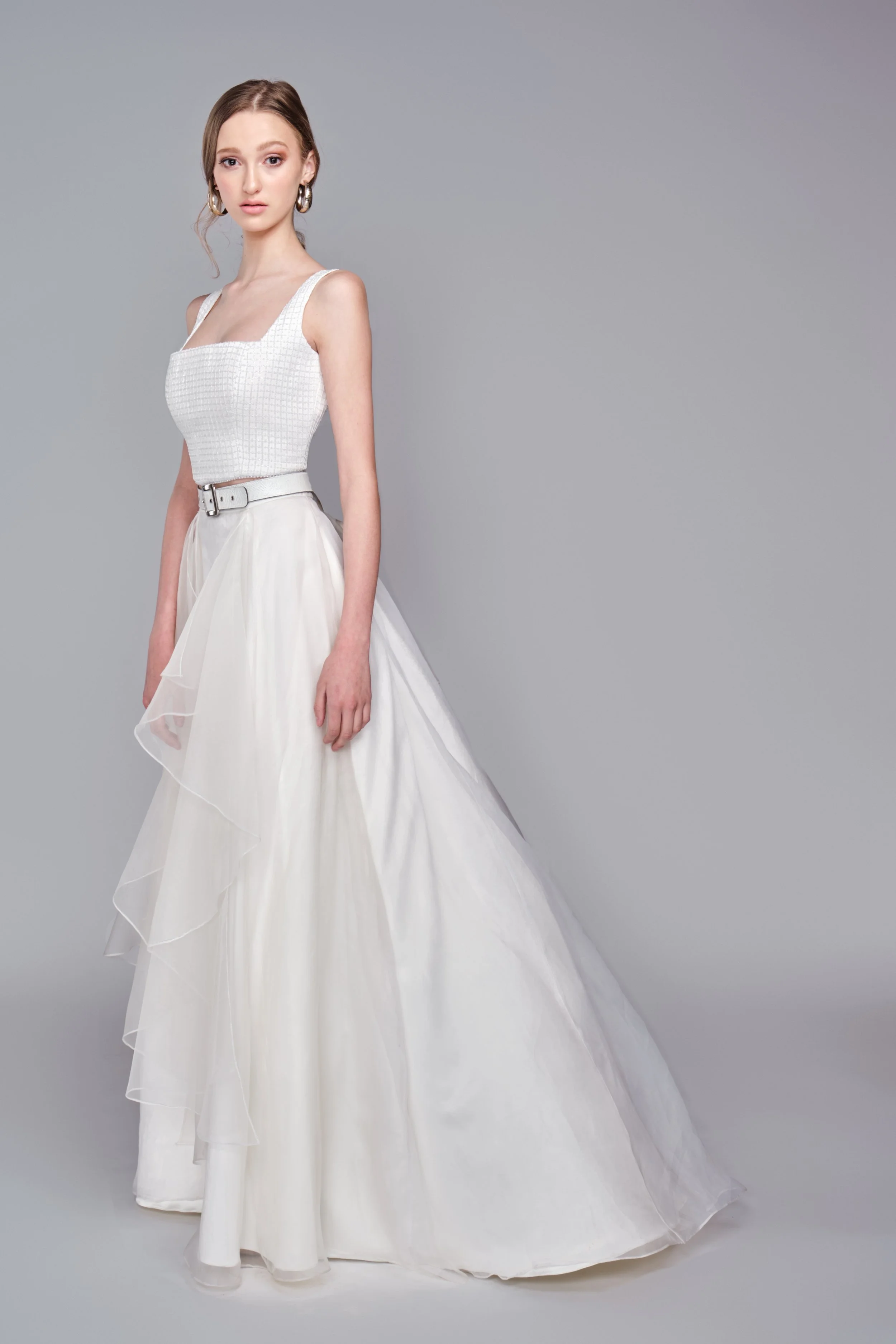Woman wearing a white sleeveless gown with a square neckline and tiered skirt, standing against a plain gray background.
