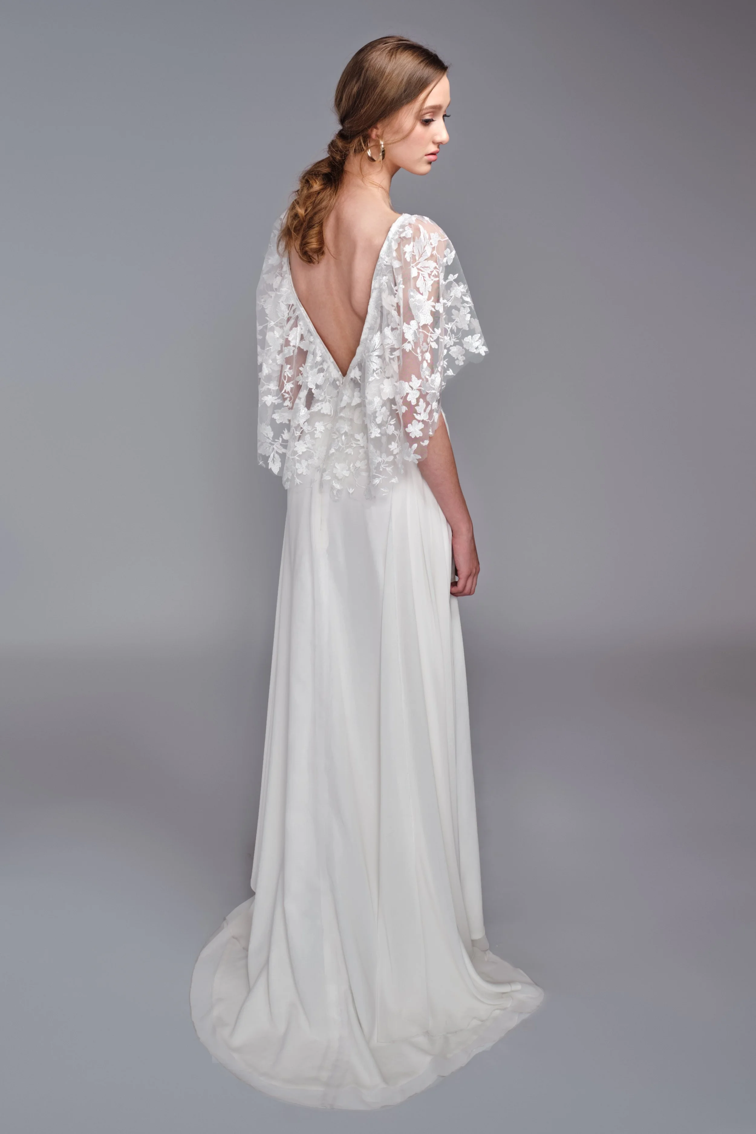 A young woman with wavy brown hair in a side profile wearing a white dress with a deep V back, lace floral overlay on the upper part, and long flowing skirt, standing against a plain gray background.