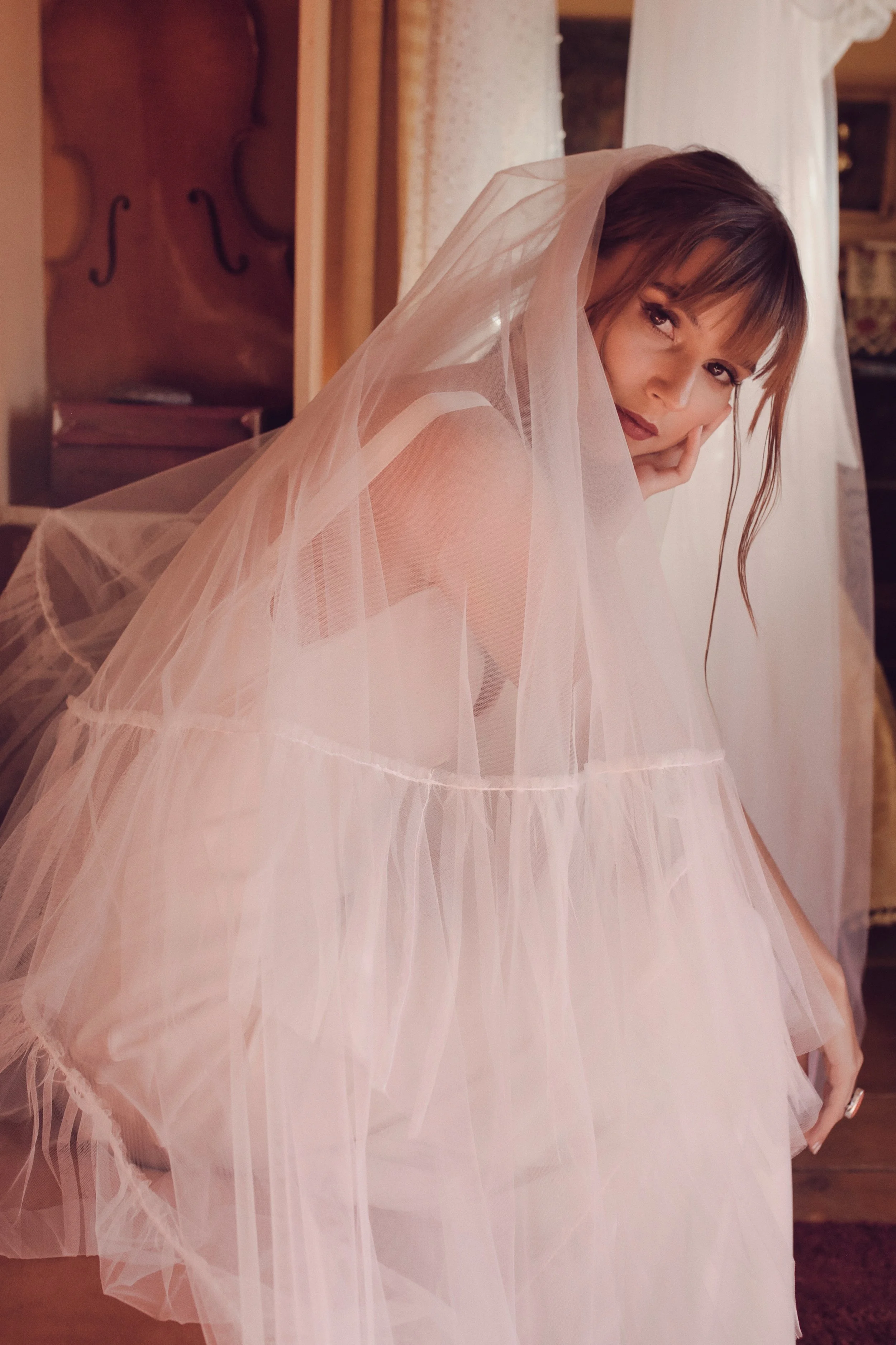 A woman with long brown hair and bangs, wearing a light pink or peach gown and veil, leaning on a wooden surface in a warmly lit room.