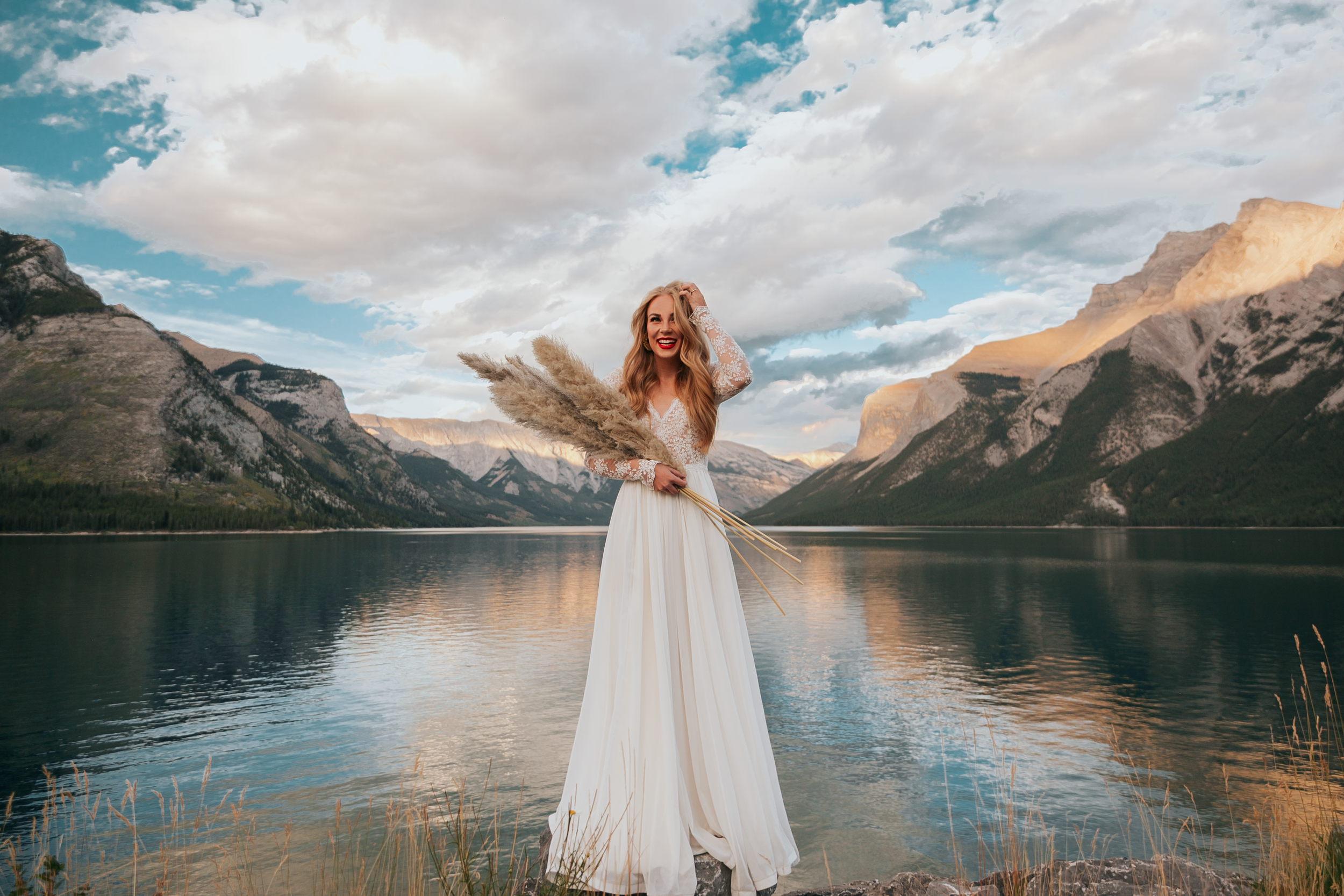 A woman in a white dress holding a large bouquet of pampas grass stands near a lake with mountains in the background and a cloudy sky overhead.