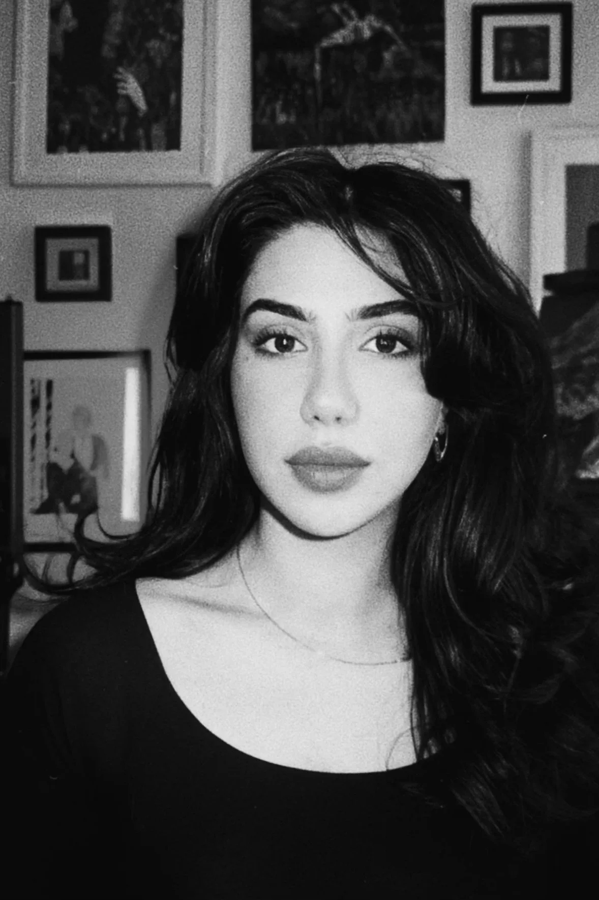 Black and white portrait of a woman with long wavy hair, wearing a dark top, with framed pictures on the wall behind her.