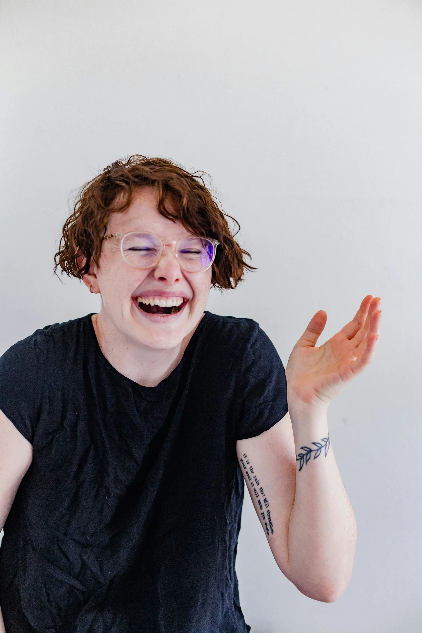 Woman with short curly red hair and glasses, laughing with eyes closed, wearing a black shirt, and gesturing with her right hand, against a plain light-colored wall.