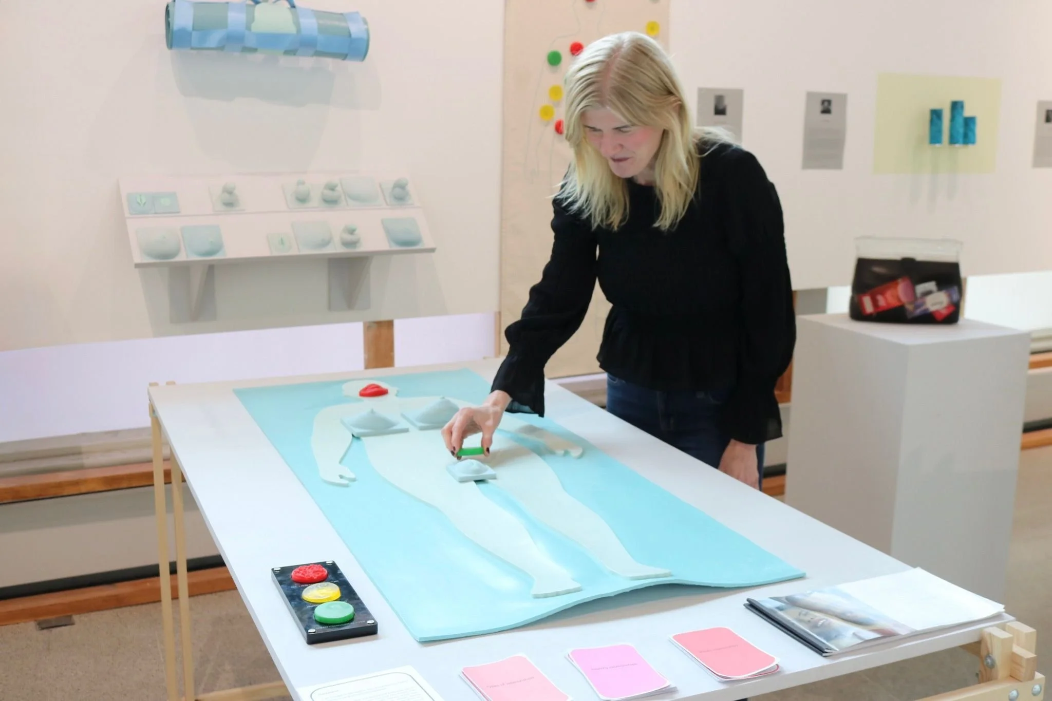 A woman with long blonde hair, wearing a black top, is leaning over a table, demonstrating one of Included's 3D learning tools, a body mat, which helps adults with disabilities explore consent.