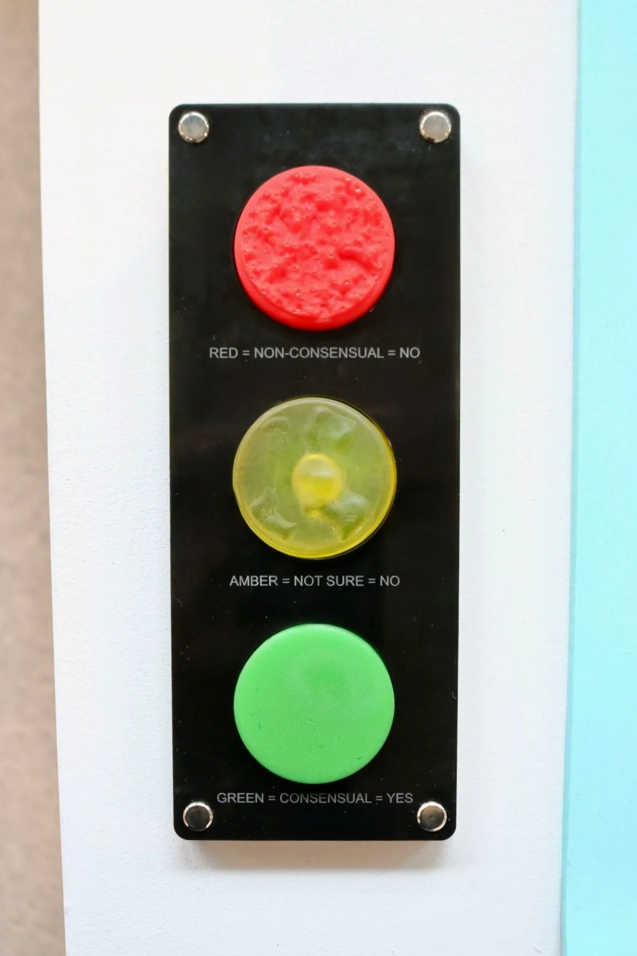Three vertically arranged buttons on a black panel, each with labels indicating their functions: a red button labeled 'RED = NON-CONSENSUAL = NO,' a yellow button labeled 'AMBER = NOT SURE = NO,' and a green button labeled 'GREEN = CONSENSUAL = YES.'