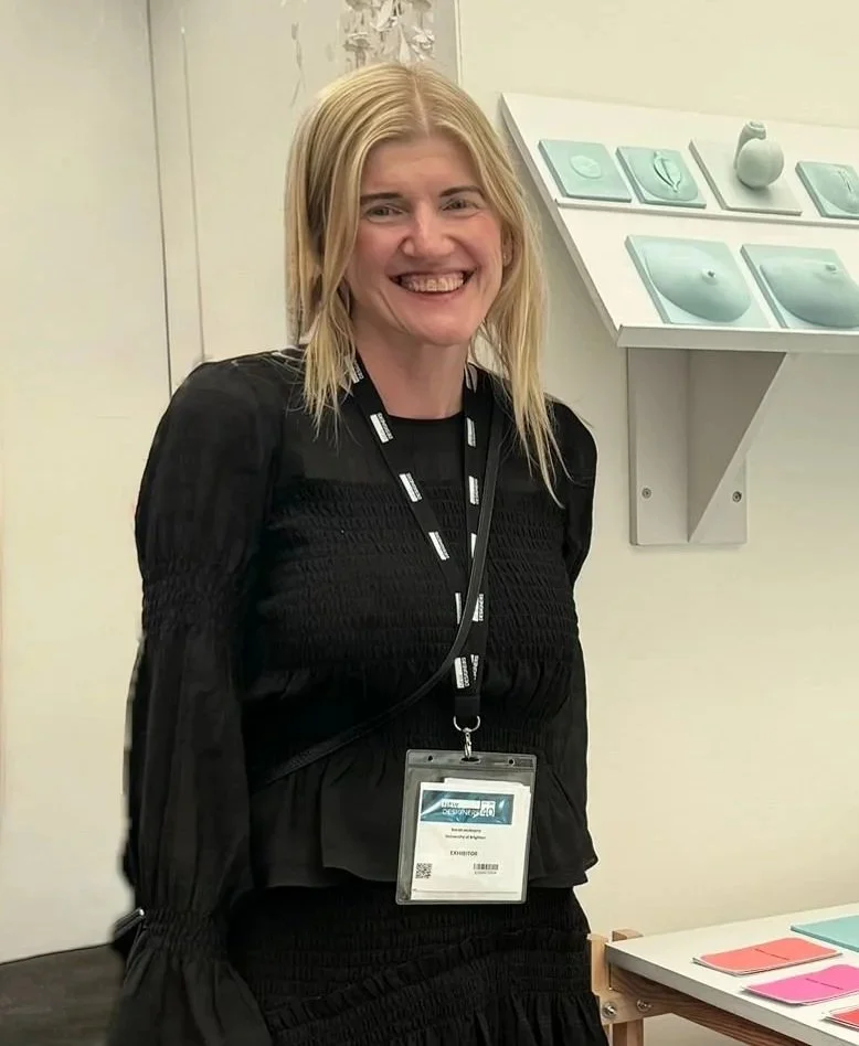 A smiling woman with blonde hair, our founder Sarah, is wearing a black top and badge on a lanyard, standing indoors near a display of Included's 3D learning tools.