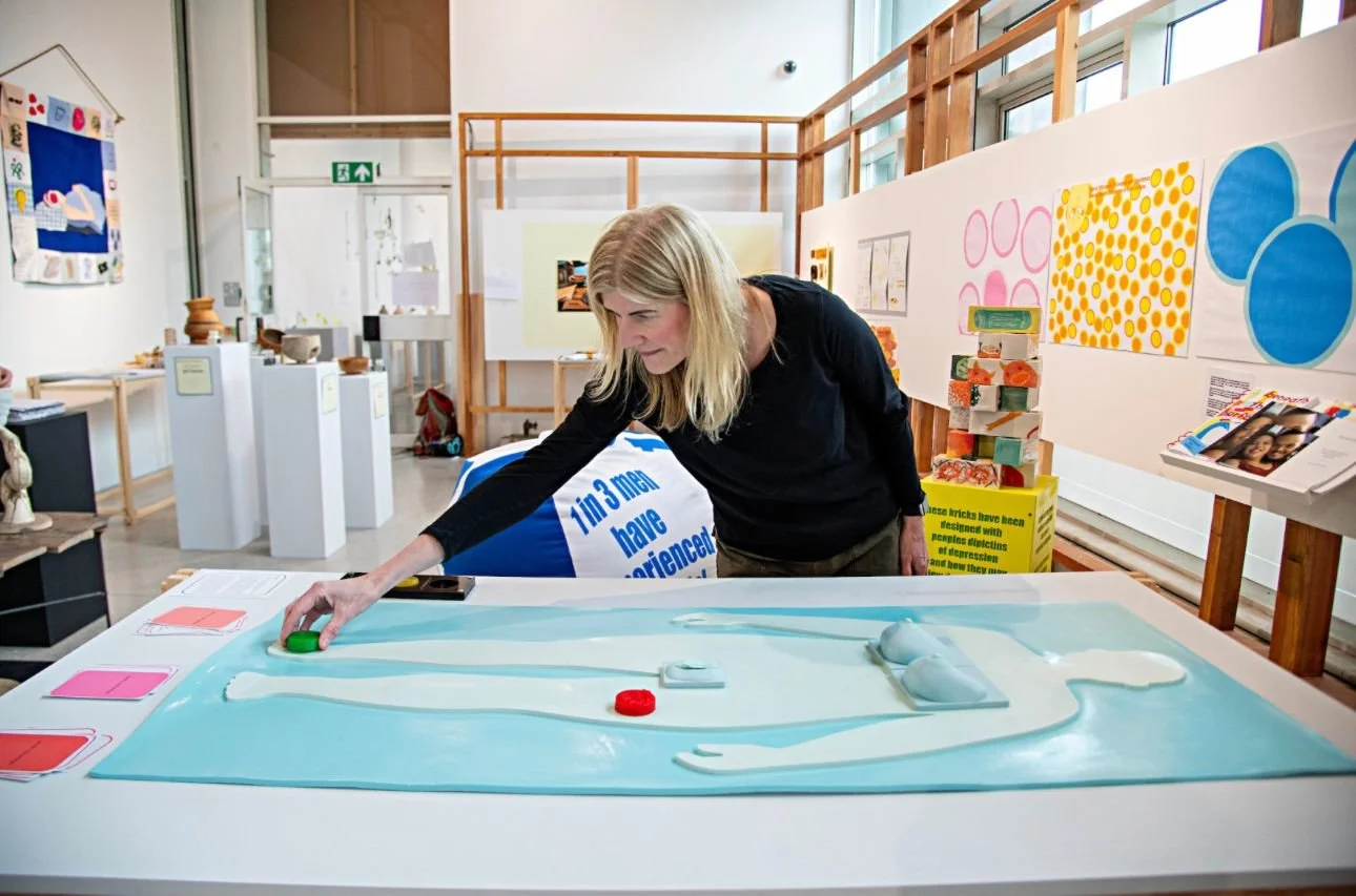 A woman with blonde hair and a black top is using one of Included's 3D learning tools to deliver a workshop in a brightly lit room decorated with artwork and informational displays.