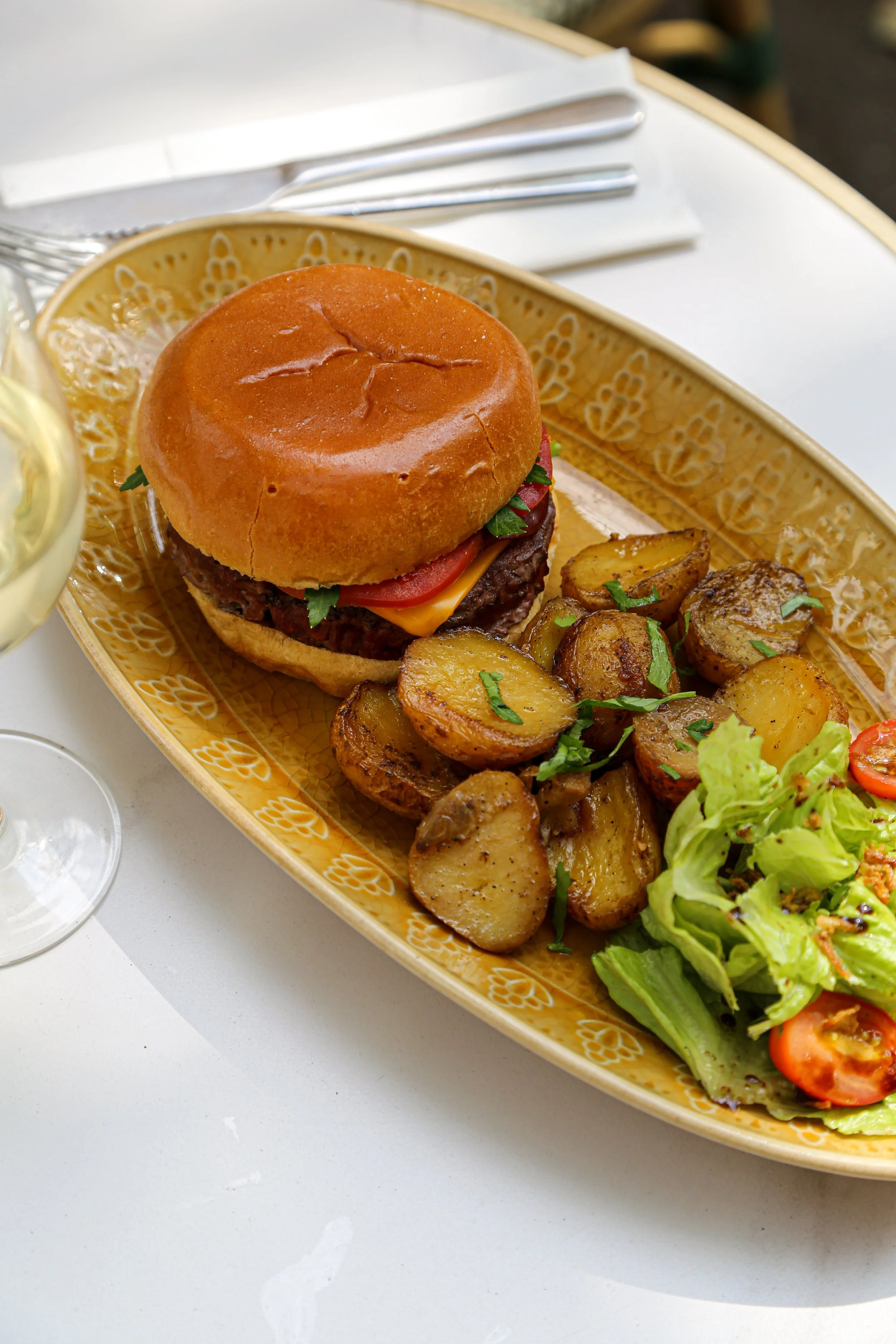 Un plateau avec un burger contenant un bun doré, une tranche de tomate, du fromage, un steak haché, des pommes de terre rôties, une salade verte et des tomates cerises.