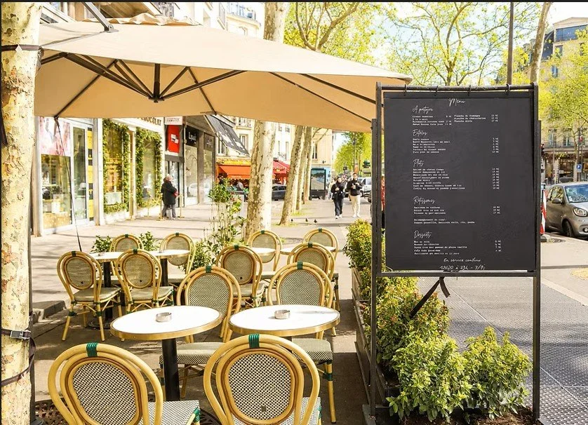 Terrasse d'un café en plein air avec des chaises en rotin et des tables, sous un grand parasol, sur un trottoir urbain avec des arbres et des bâtiments en arrière-plan.
