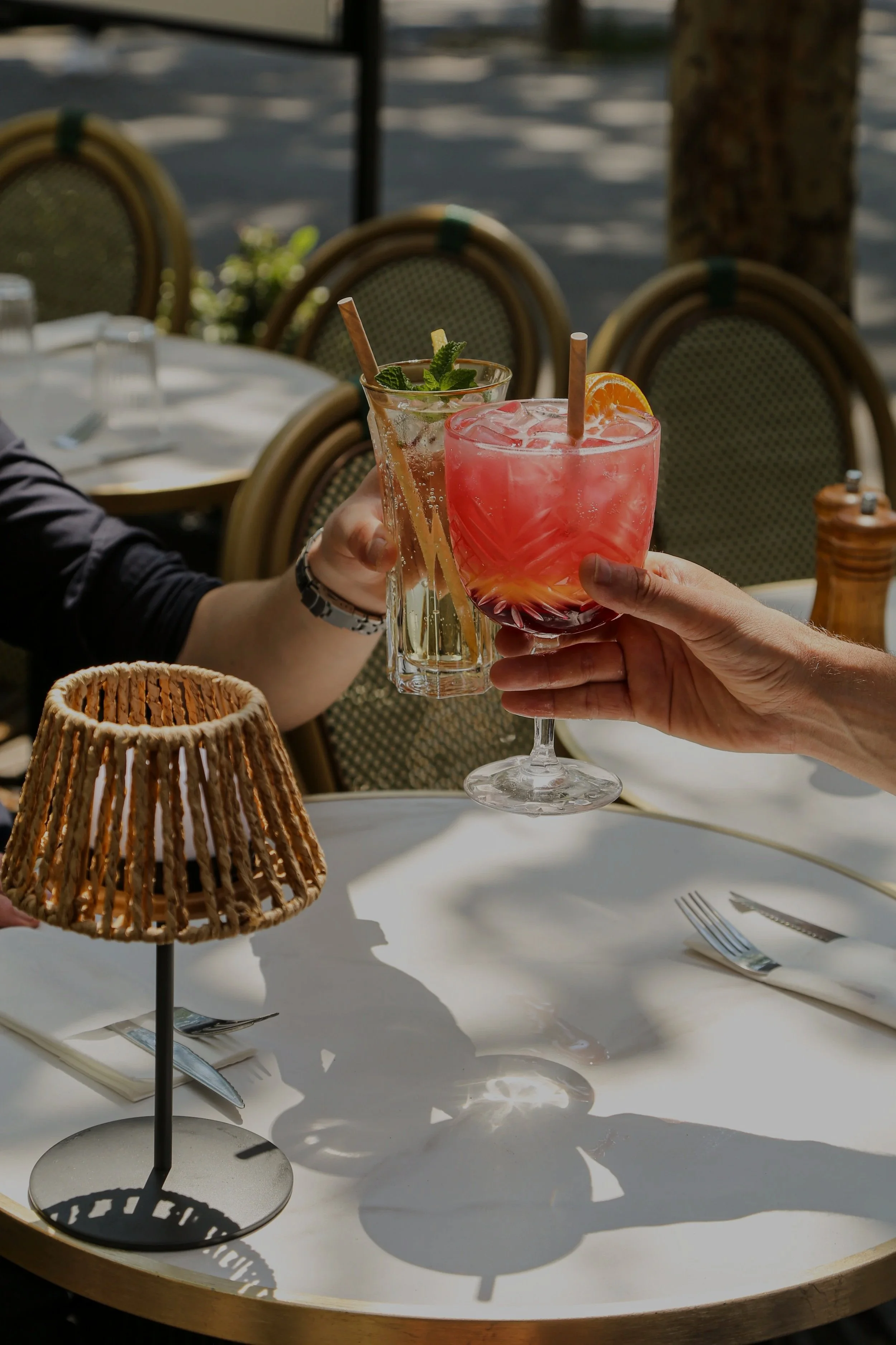 Deux cocktails colorés avec glaçons, une tranche d'orange et de la menthe, tenus par deux personnes. Décor de terrasse de restaurant avec des chaises en rotin et une lampe en osier.