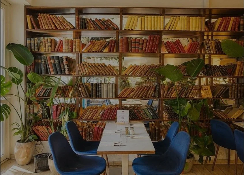 Une salle à manger avec une table en bois, entourée de six chaises en velours bleu foncé. La table est prête pour un repas, avec des couverts, des verres et une petite enseigne. En fond, un immense rayonnage en bois rempli de livres anciens et de couleurs variées, encadré par des grandes plantes vertes.