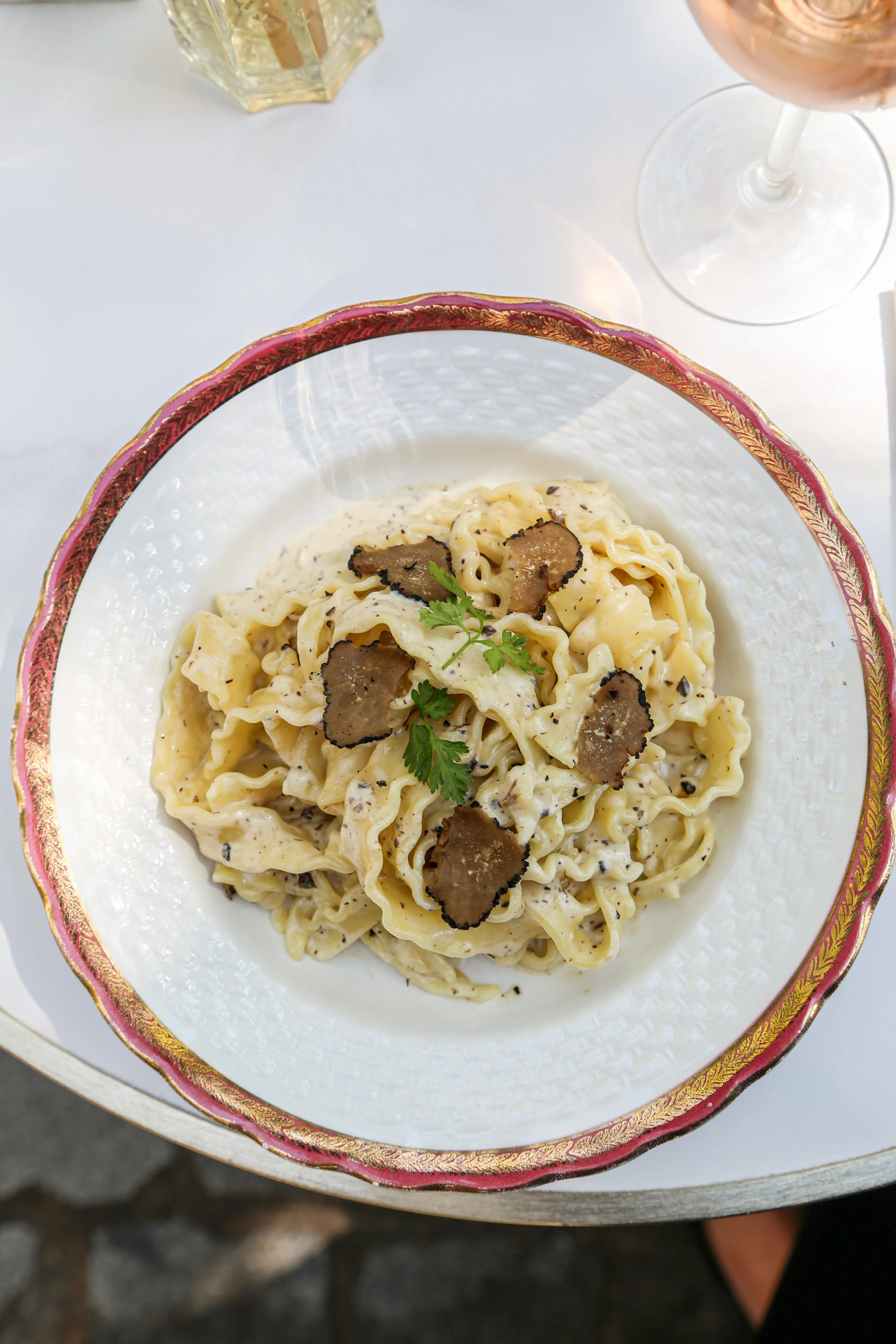 Plat de pâtes aux truffes avec crème, décoré de persil, servi dans une assiette élégante avec une bordure dorée et rose, placé sur une table blanche.