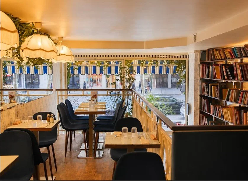 Intérieur d'une café ou bibliothèque avec tables, chaises, étagères de livres, grande fenêtre avec vue sur la rue