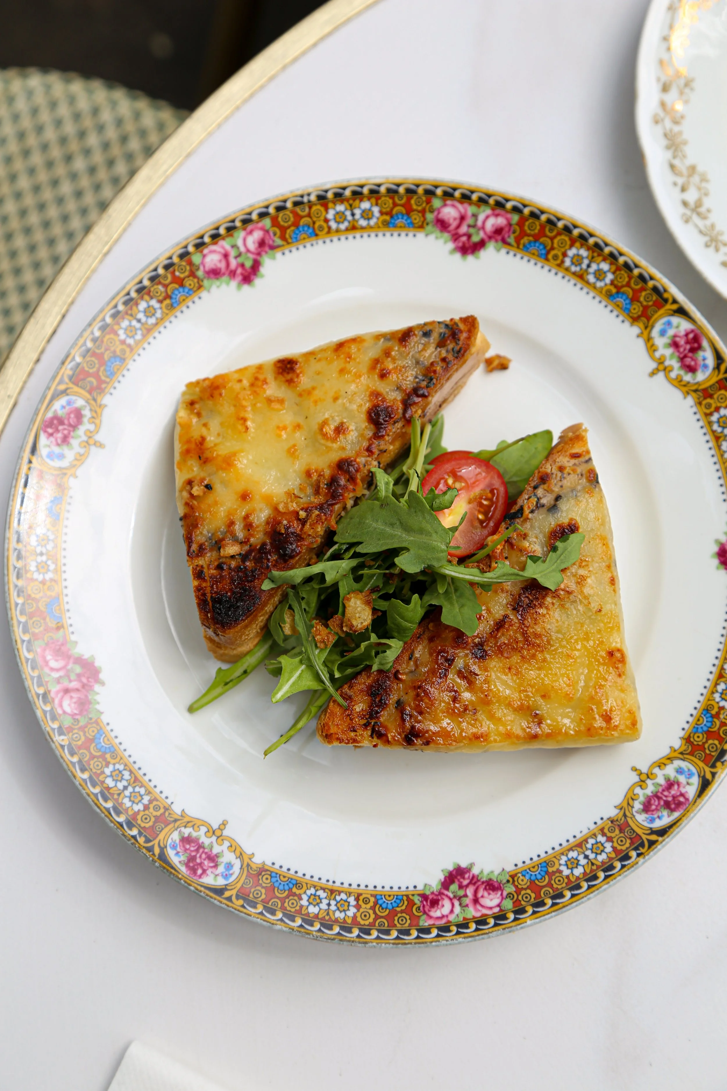 Une assiette de pizza avec du fromage fondu, garnie de roquette, tomate et herbes sur une assiette décorative avec des motifs floraux.