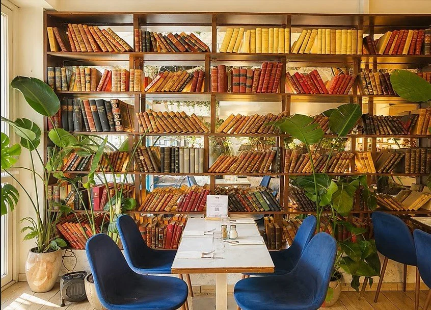 Une bibliothèque avec une étagère pleine de livres anciens, entourée de plantes vertes et un coin repas avec une table blanche et des chaises bleues.