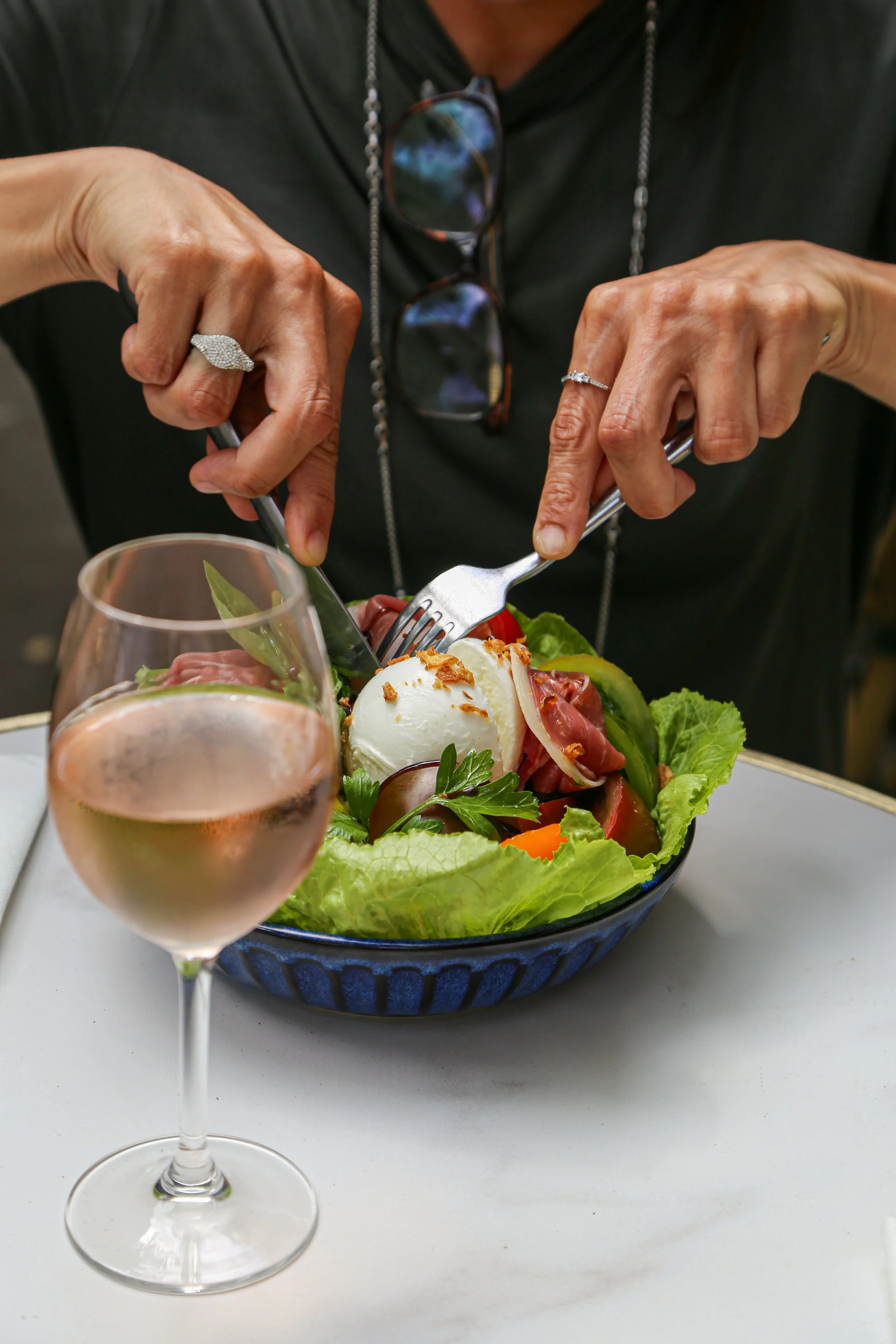 Une personne mange une salade à l'aide d'une fourchette, en présence d'un verre de vin rosé. La salade est composée de divers légumes et de mozzarella, dans un bol bleu.