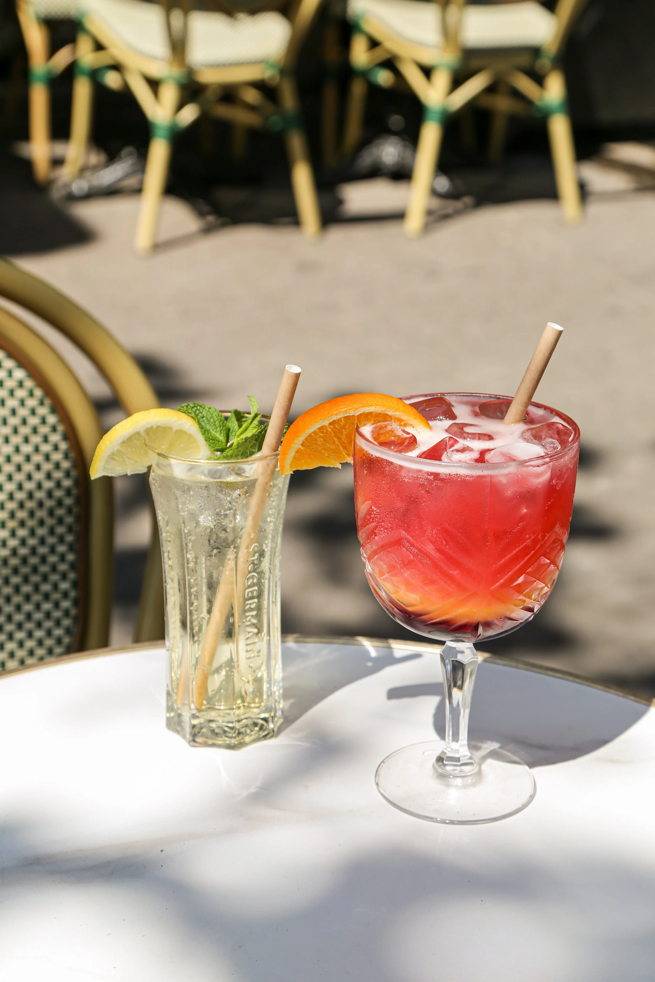 Deux cocktails en verre, un avec une tranche d'orange et un autre avec une tranche de citron, garnis de feuilles de menthe, sur une table blanche sous un soleil brillant, avec des chaises en rotin en arrière-plan.