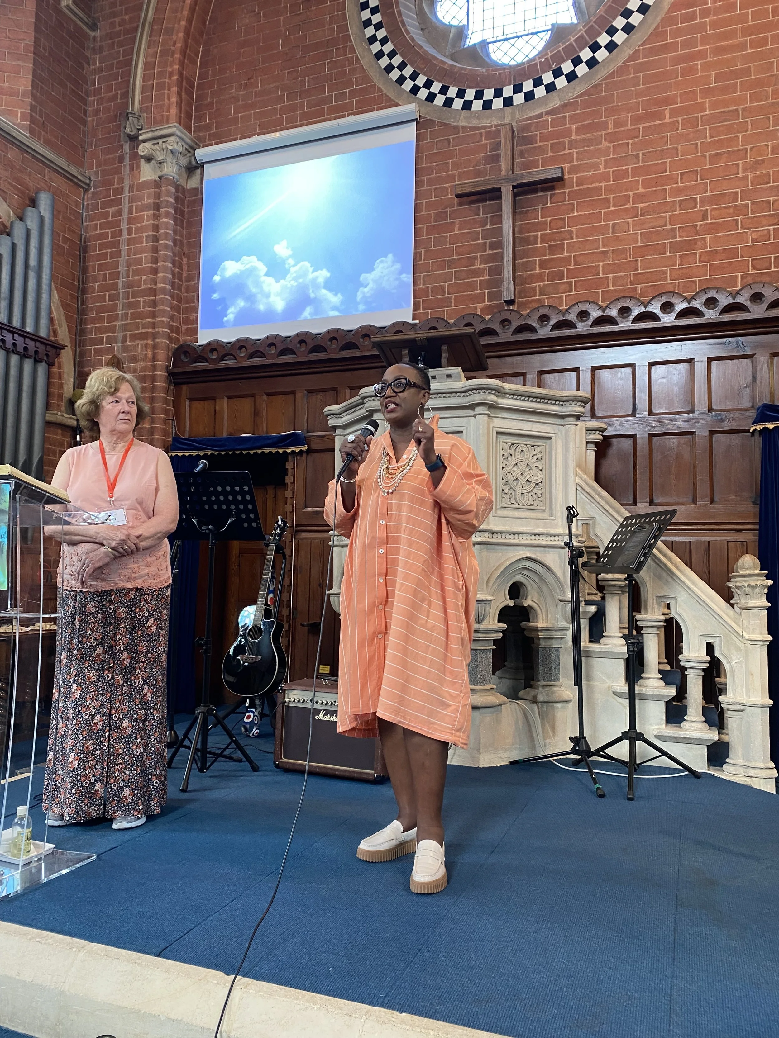 A woman speaking into a microphone at a church. She is wearing glasses, a peach striped dress, and white shoes. Another woman stands nearby. The church interior has red brick walls, a cross, a stained-glass window, and musical instruments.