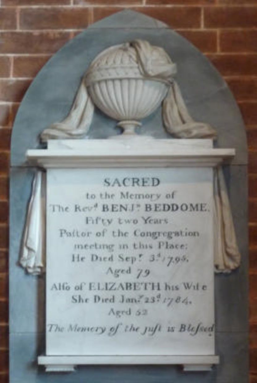 A memorial plaque with a decorative urn and drapes carved in relief above it. The plaque is dedicated to Reverend Benjamin Beddome, mentioning his death on September 3rd, 1795, at the age of 79, and his wife Elizabeth, who died on January 23rd, 1784, at the age of 62.