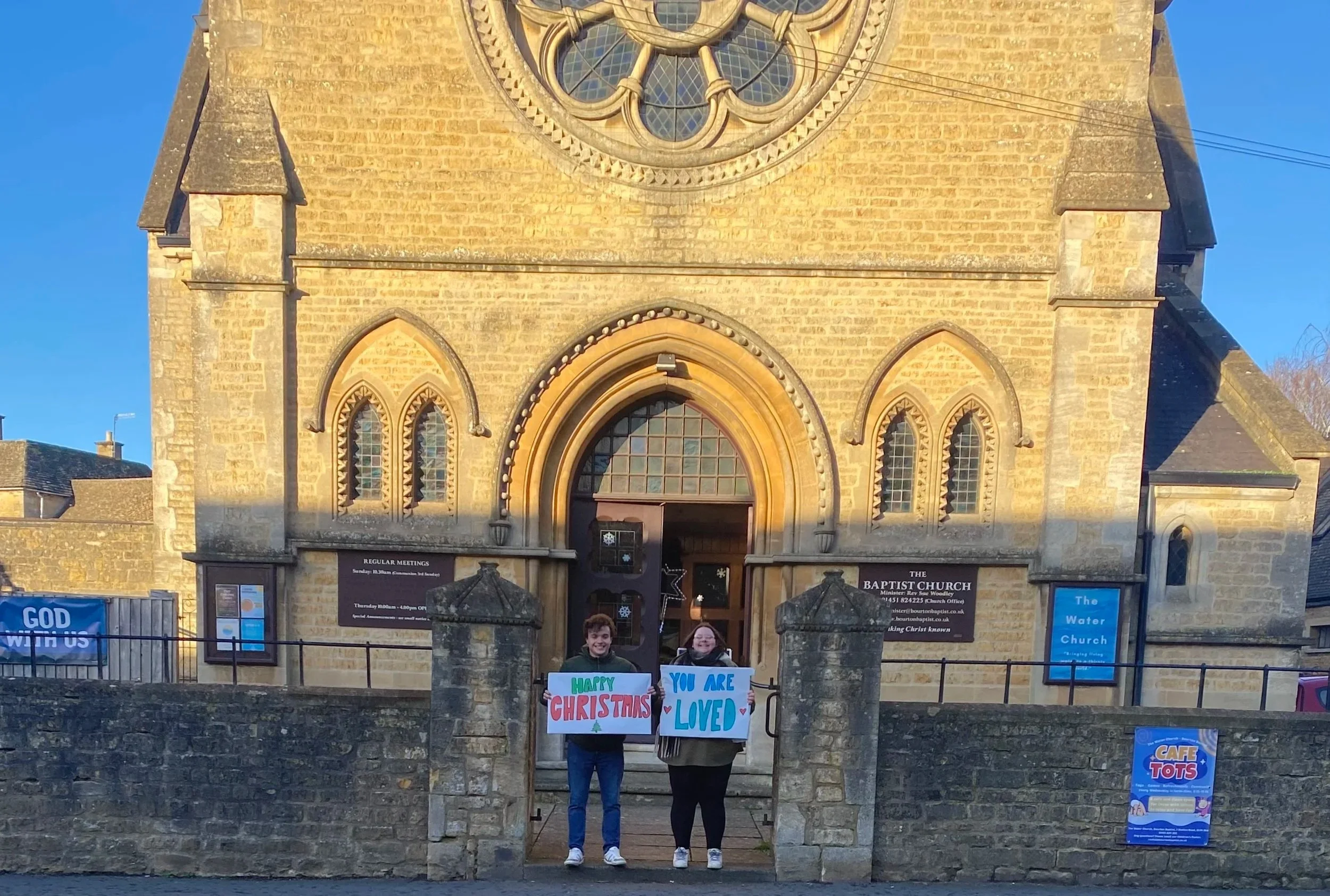 People standing in front of the Baptist Church holding Christmas signs that say "Merry Christmas" and "You are loved." The church has a large circular stained glass window above the entrance and stone architecture.