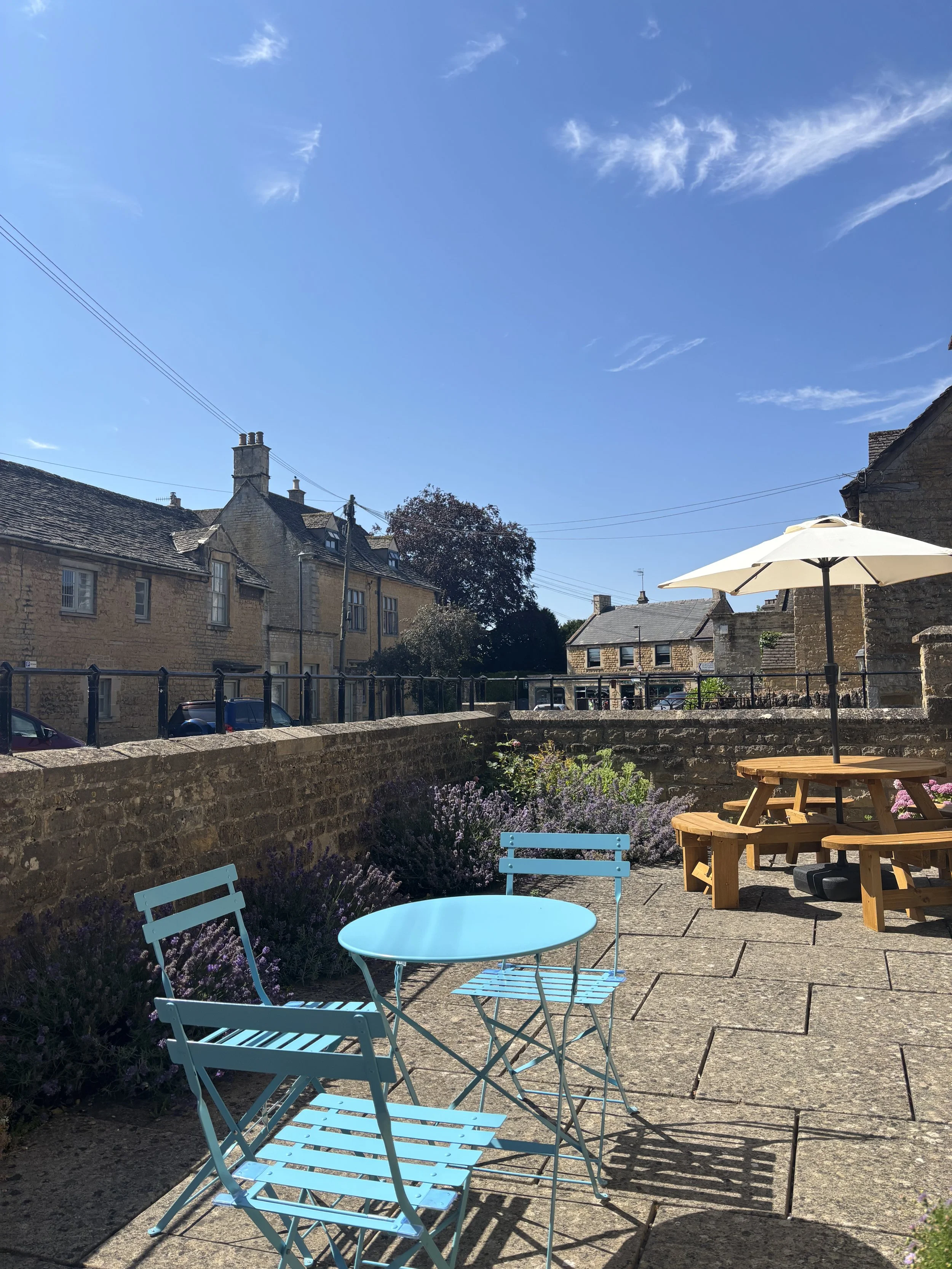A sunny outdoor seating area with a round blue table, matching chairs, picnic tables, a large umbrella, and purple flowering plants in front of stone buildings and a blue sky with clouds.