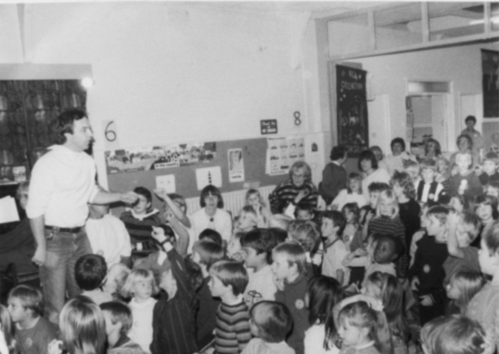 A black-and-white photo of a group of children sitting and standing in an indoor space, with a man handing something to a child, in a setting resembling a school or community center, with posters and signs visible on the walls.