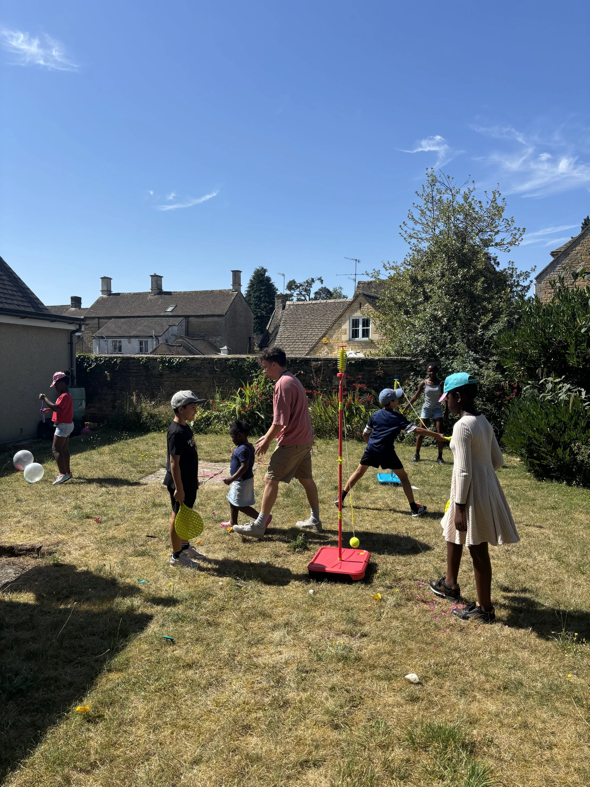 Children and adults playing a game of outdoor tennis or a similar sport in a backyard on a sunny day.