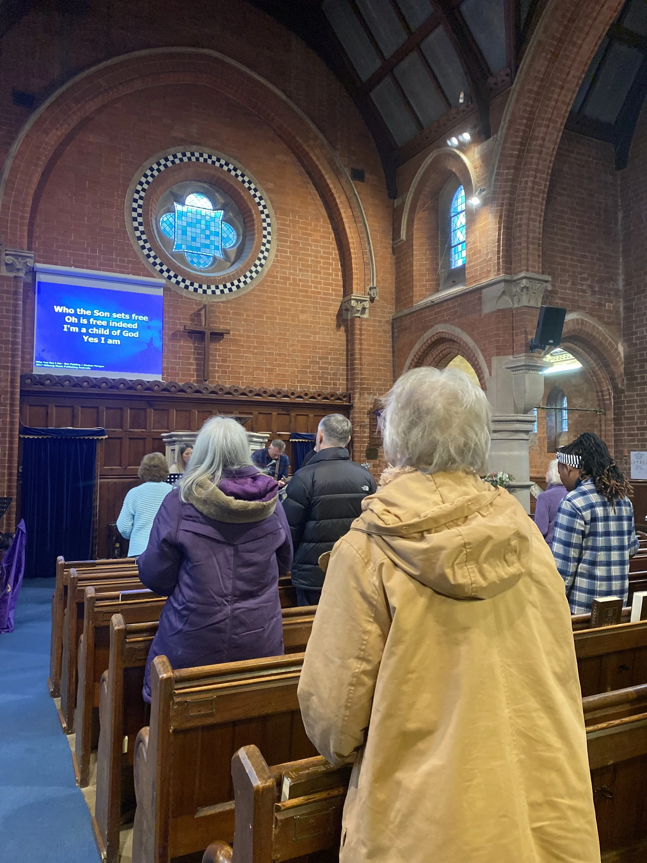 Inside a church, people are gathered for a service. The church has brick walls, stained glass windows, and wooden pews. There is a screen displaying lyrics of a hymn or song.