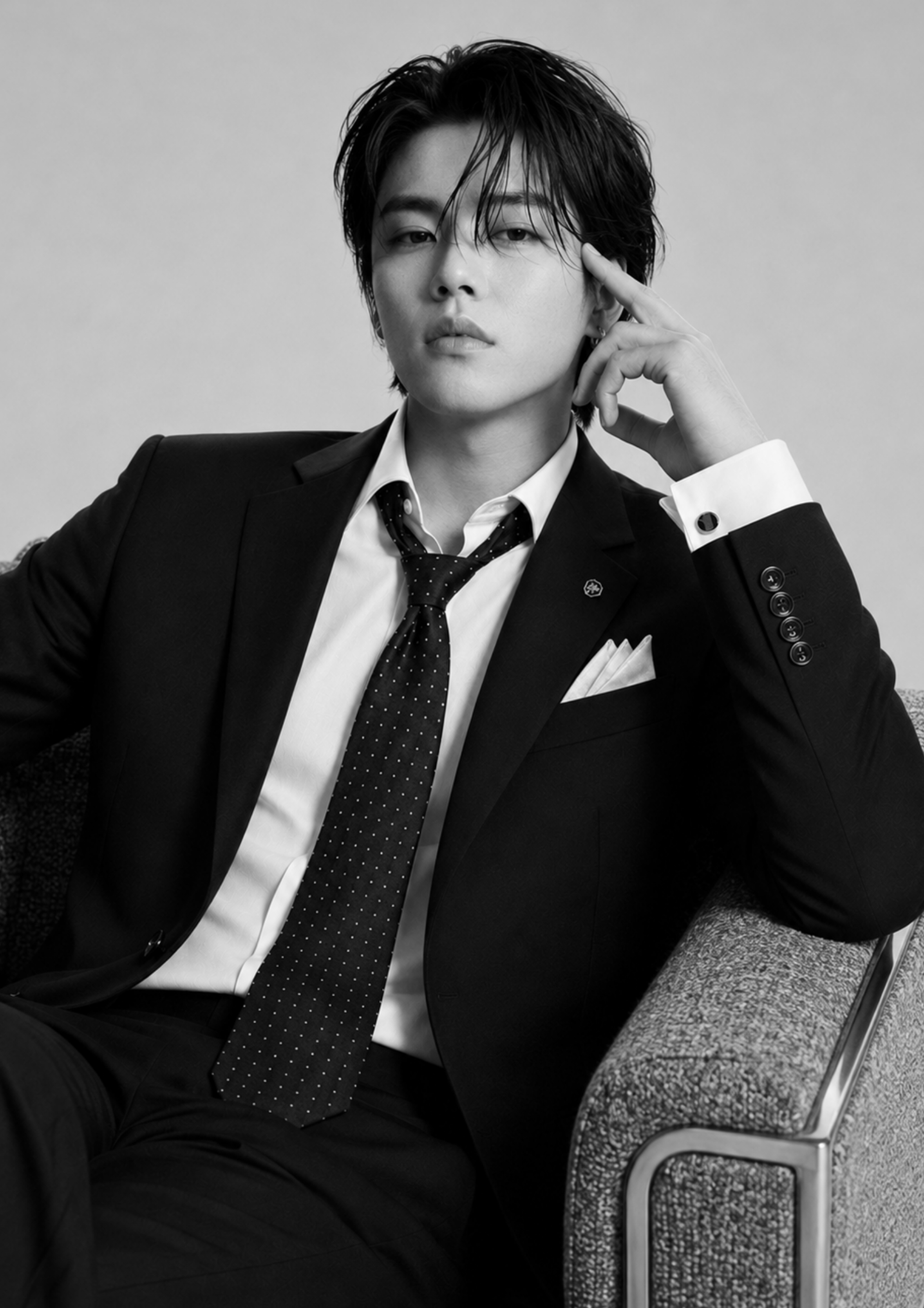 Hombre con cabello oscuro y peinado desordenado, vestimenta formal con saco y corbata, sentado en una silla con respaldo de tela, mirando hacia la cámara en un entorno de interior, en blanco y negro.