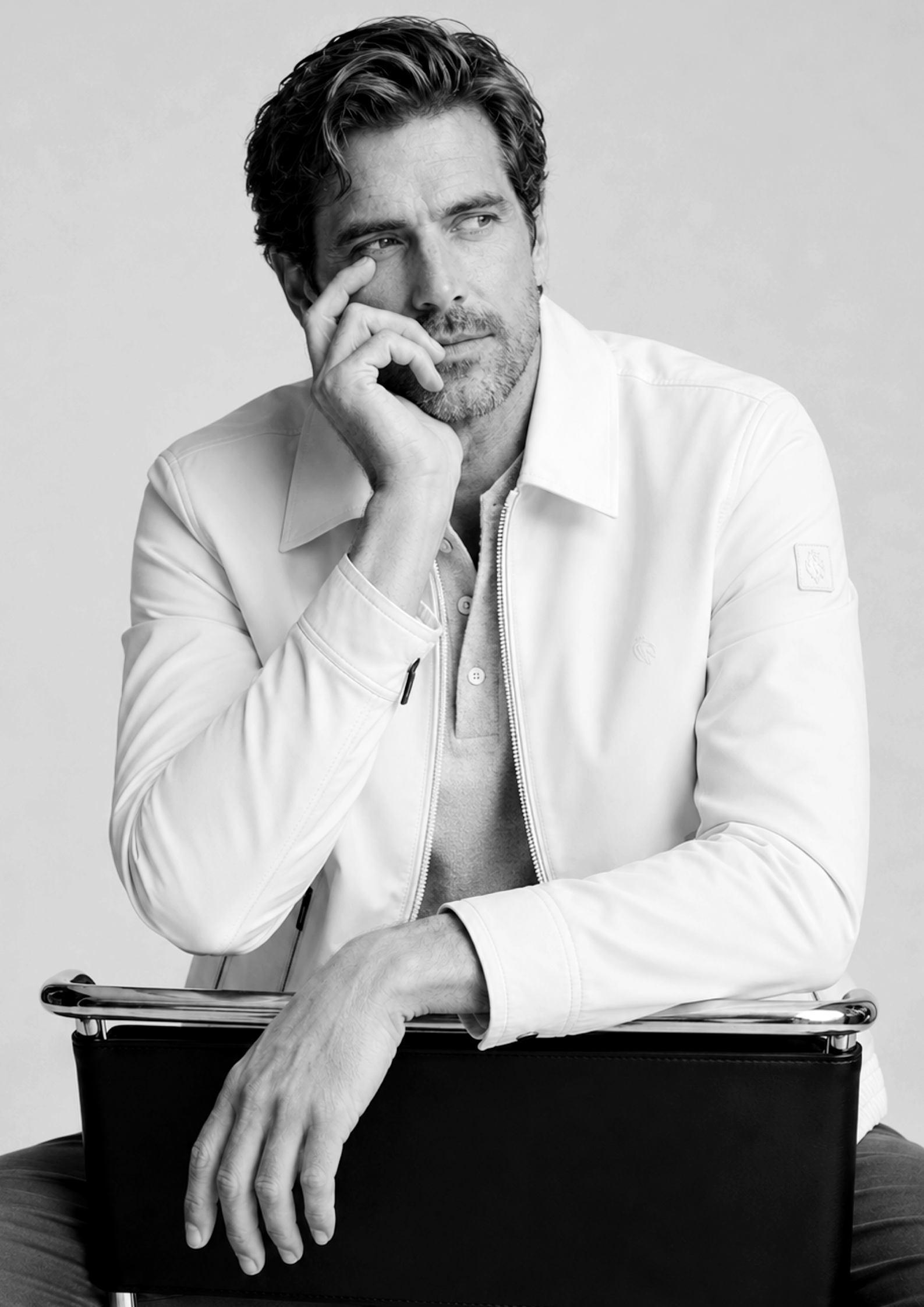 Hombre con barba y cabello rizado, en blanco y negro, sentado con una mano en la cara, usando chaqueta blanca y con un dispositivo electrónico, en un fondo gris claro.