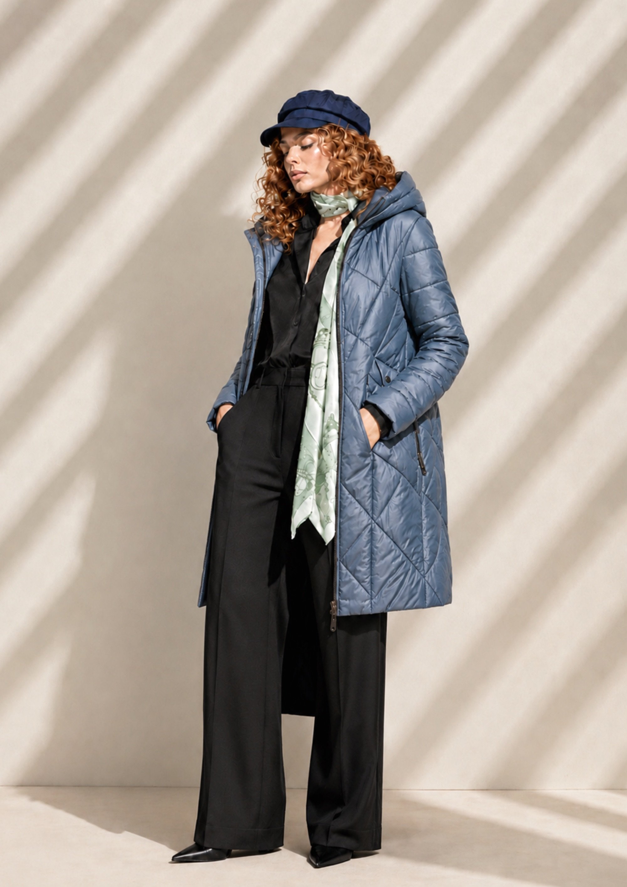 Mujer con cabellera rizada de color castaño y usar ropa de abrigo, incluyendo un abrigo azul, pantalones anchos negros y sombrero azul, de pie contra fondo gris con rayas de sombra.