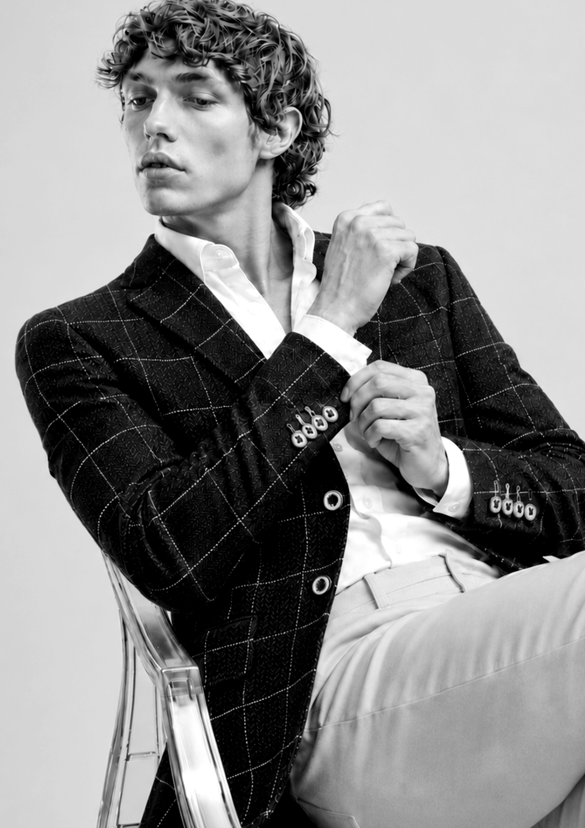 Hombre con cabello rizado, usando saco de cuadros y camisa blanca, sentado en una silla de plástico transparente, gesticulando con las manos, en una fotografía en blanco y negro.