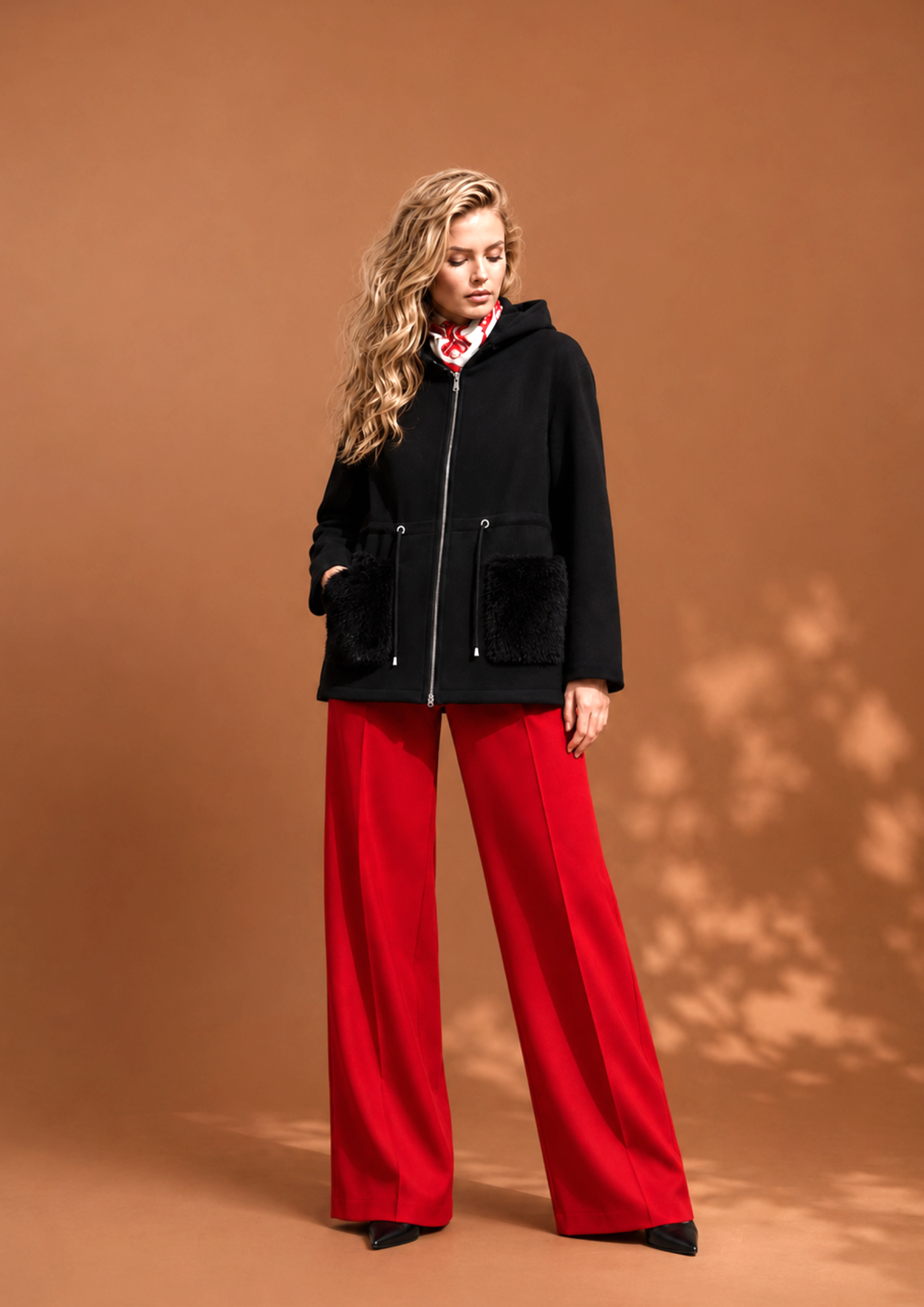 Mujer con cabello rubio rizado, usando un abrigo negro con bolsillas de peluche, pantalones anchos rojos y botas negras, posando en un fondo de color marrón