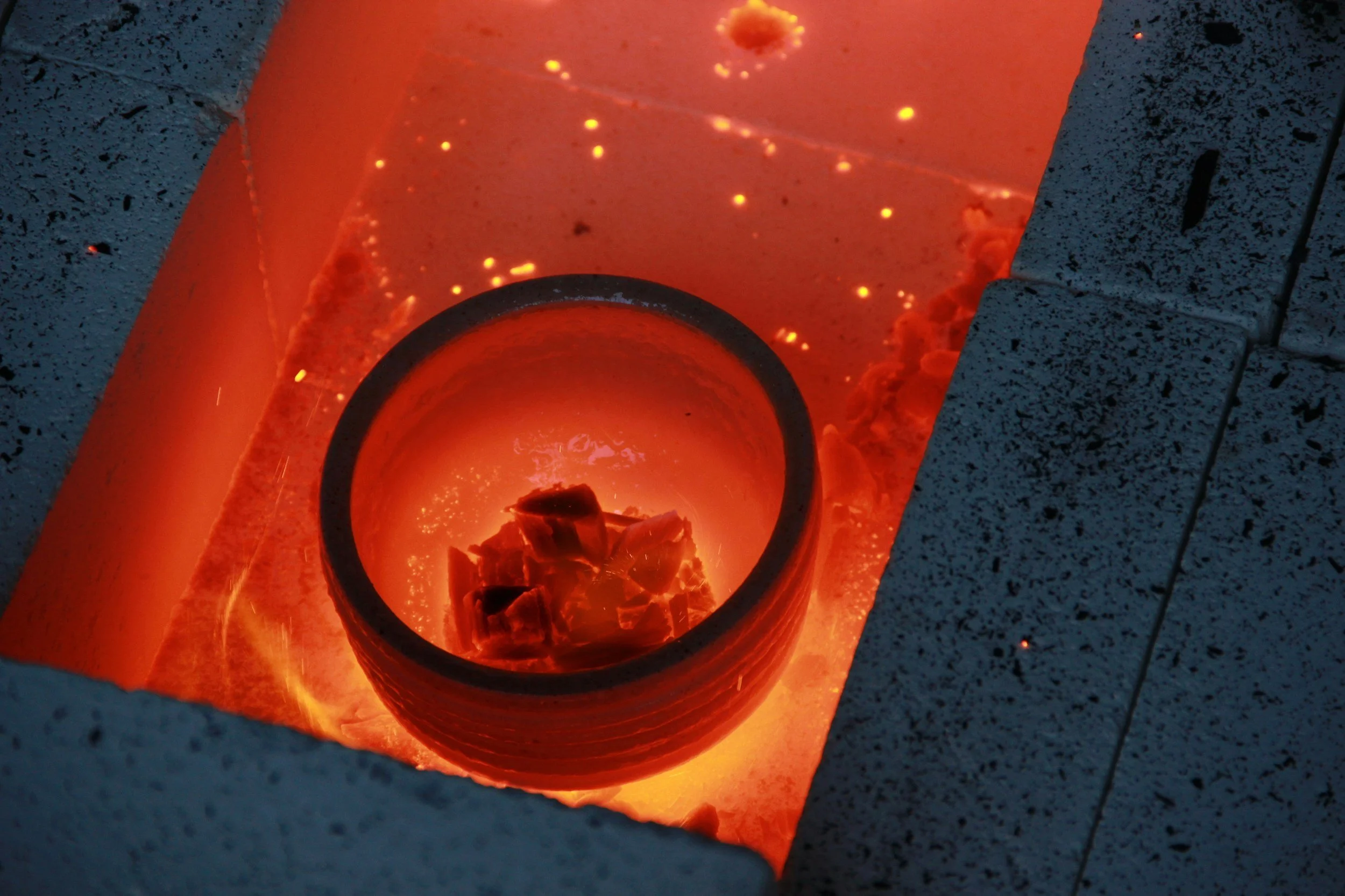 A hot glass furnace with glowing orange interior, seen through a circular opening in a stone wall, with sparks and molten material visible inside.