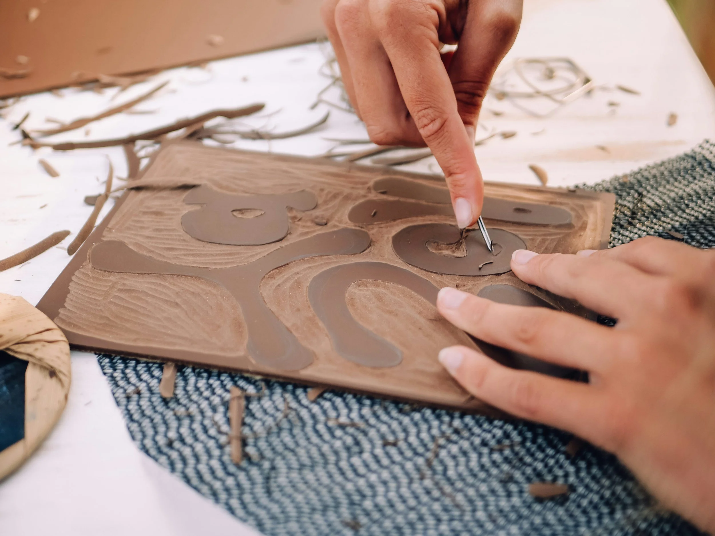 Close-up of a person's hand carving details into a wooden mold with a small metal tool, surrounded by wood shavings on a work surface.