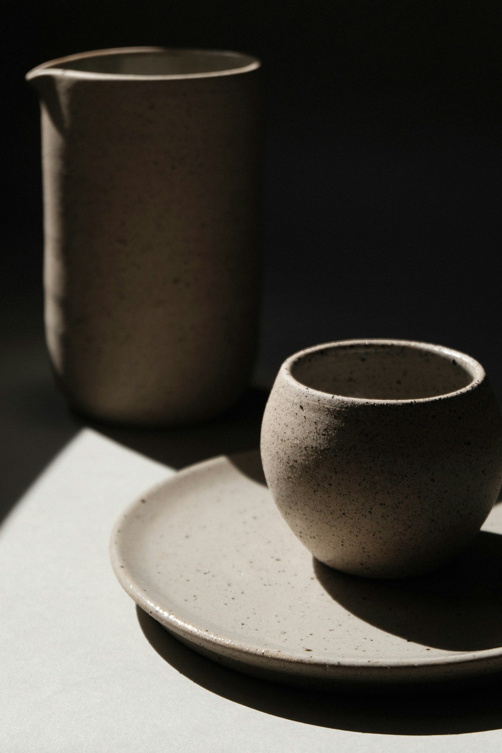 A ceramic cup and a small dish with a plate underneath them, both with a speckled beige and dark brown glaze, arranged on a white surface.