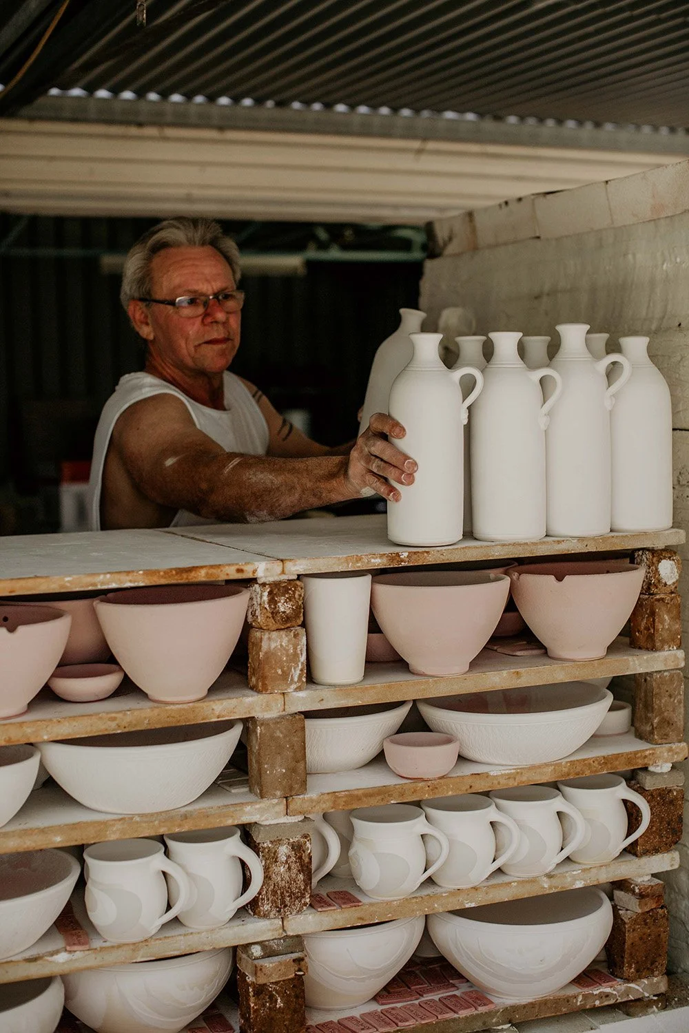 A man in a sleeveless shirt and glasses arranges white ceramic vases on a shelf in a pottery studio.