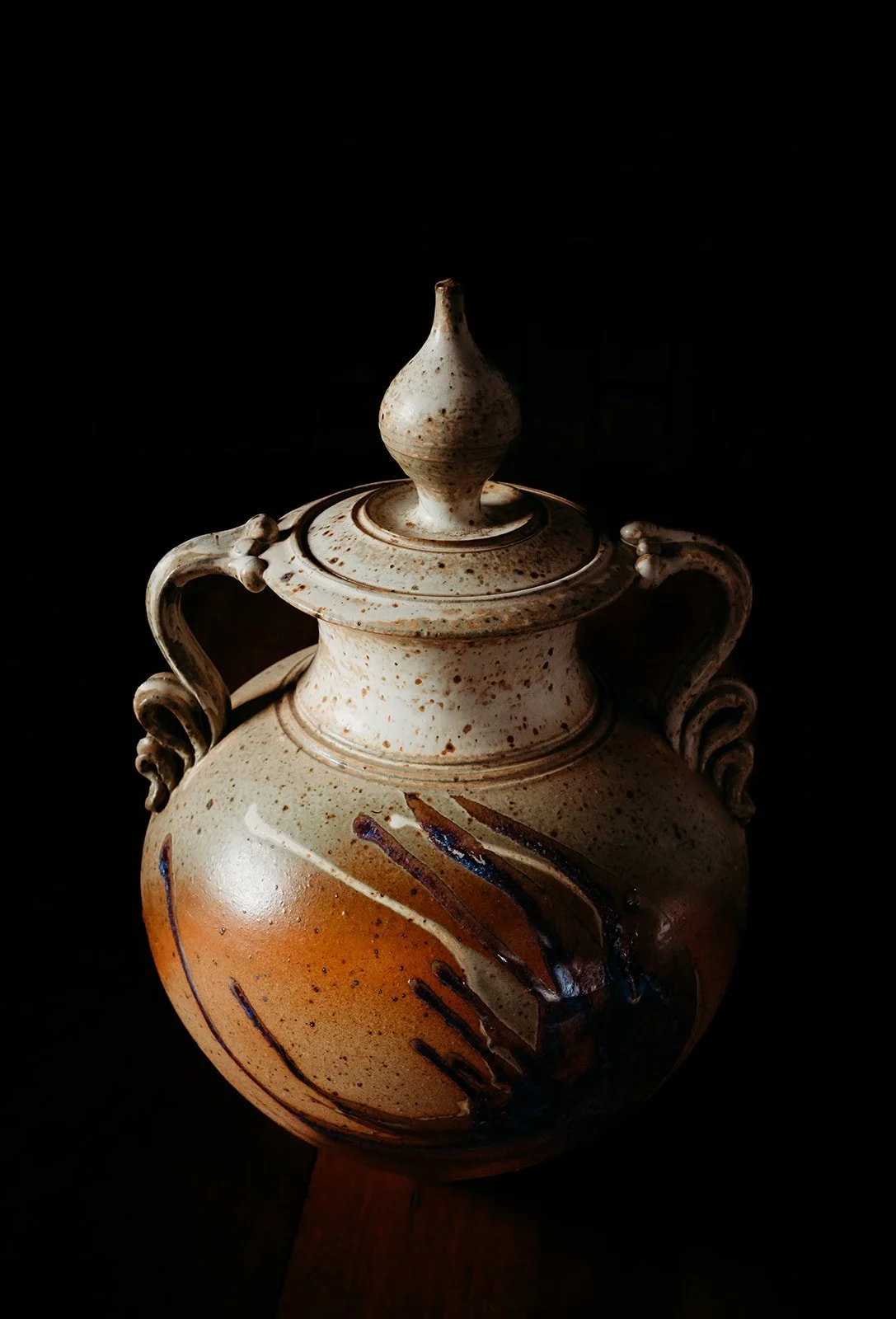 A ceramic vase with a lid and two handles, decorated with brown and black brushstrokes on a speckled beige surface, set against a dark background.