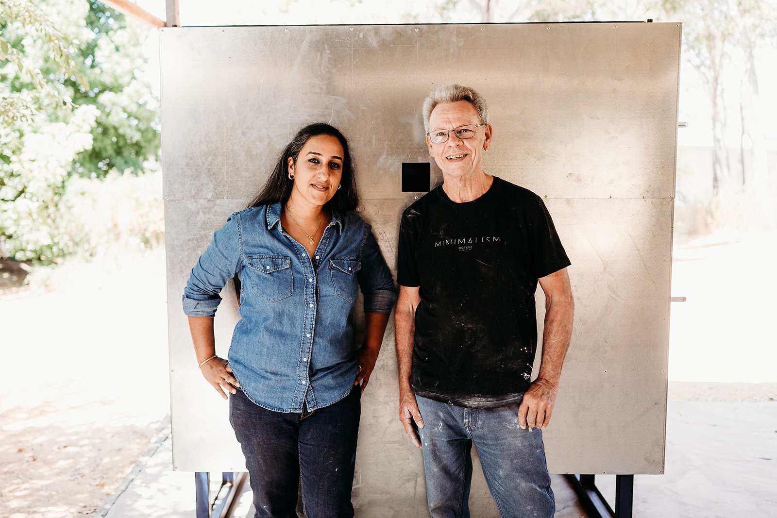 A woman in a denim shirt and a man in a black t-shirt with white paint splatters, standing outdoors in front of a large metal board, with trees visible in the background.