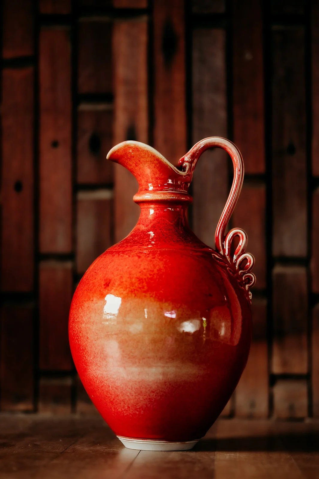 A ceramic red and orange pitcher with a curved handle and a spout, sitting on a wooden floor in front of a dark wooden backdrop.